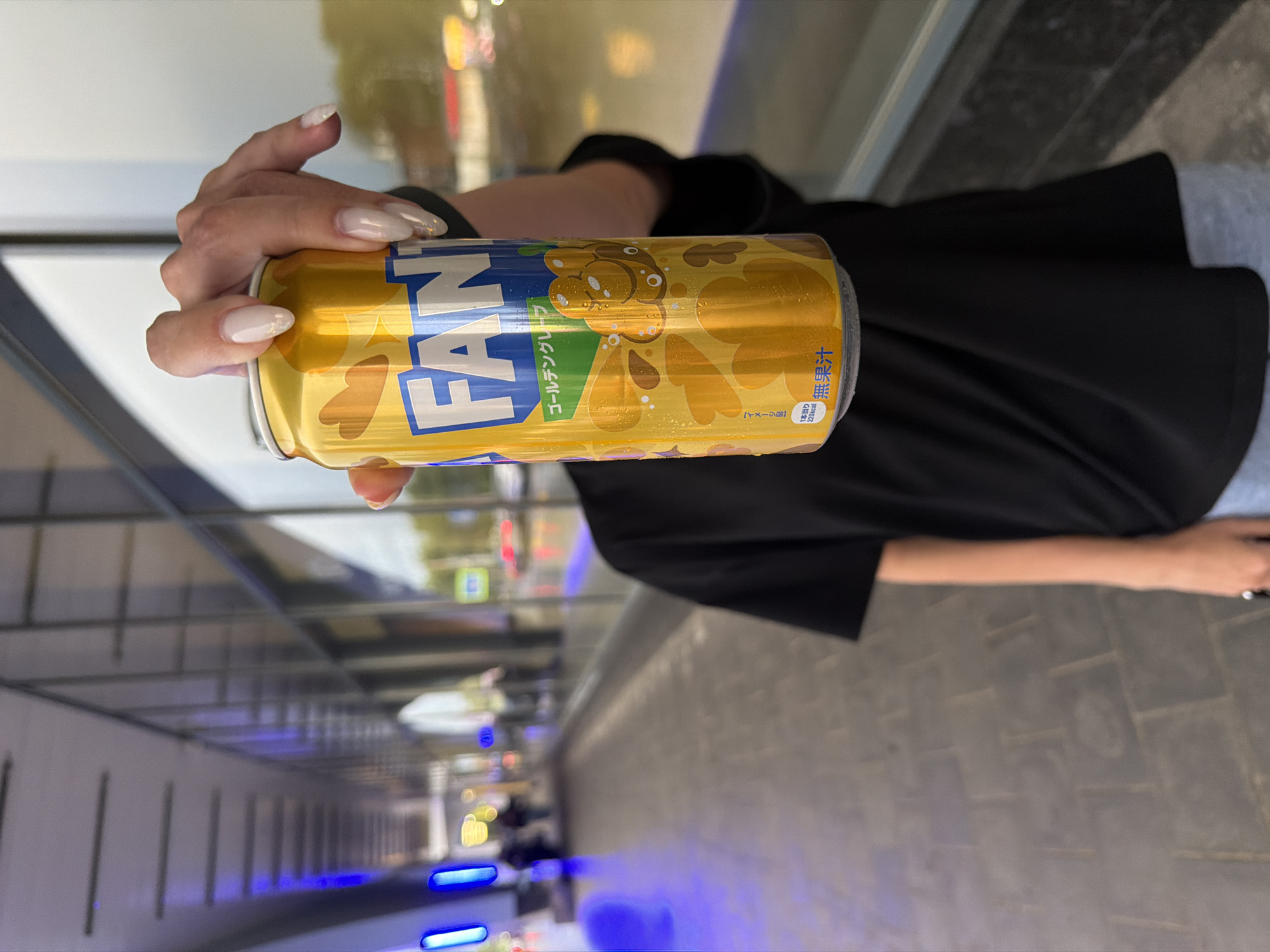A person's hand, with long, light-colored fingernails, holds a golden aluminum can. The can has blue and yellow text and graphics. The text reads "FAN" in large blue letters against a yellow background. Below this, in smaller green text, it says "ゴールデンクレフ" which is Japanese for "Golden Clef". Further text in Japanese characters is visible on the right side of the can. The background is blurred, but appears to be an outdoor setting with reflections from glass or windows, suggesting an urban environment. The lighting indicates it is daytime.