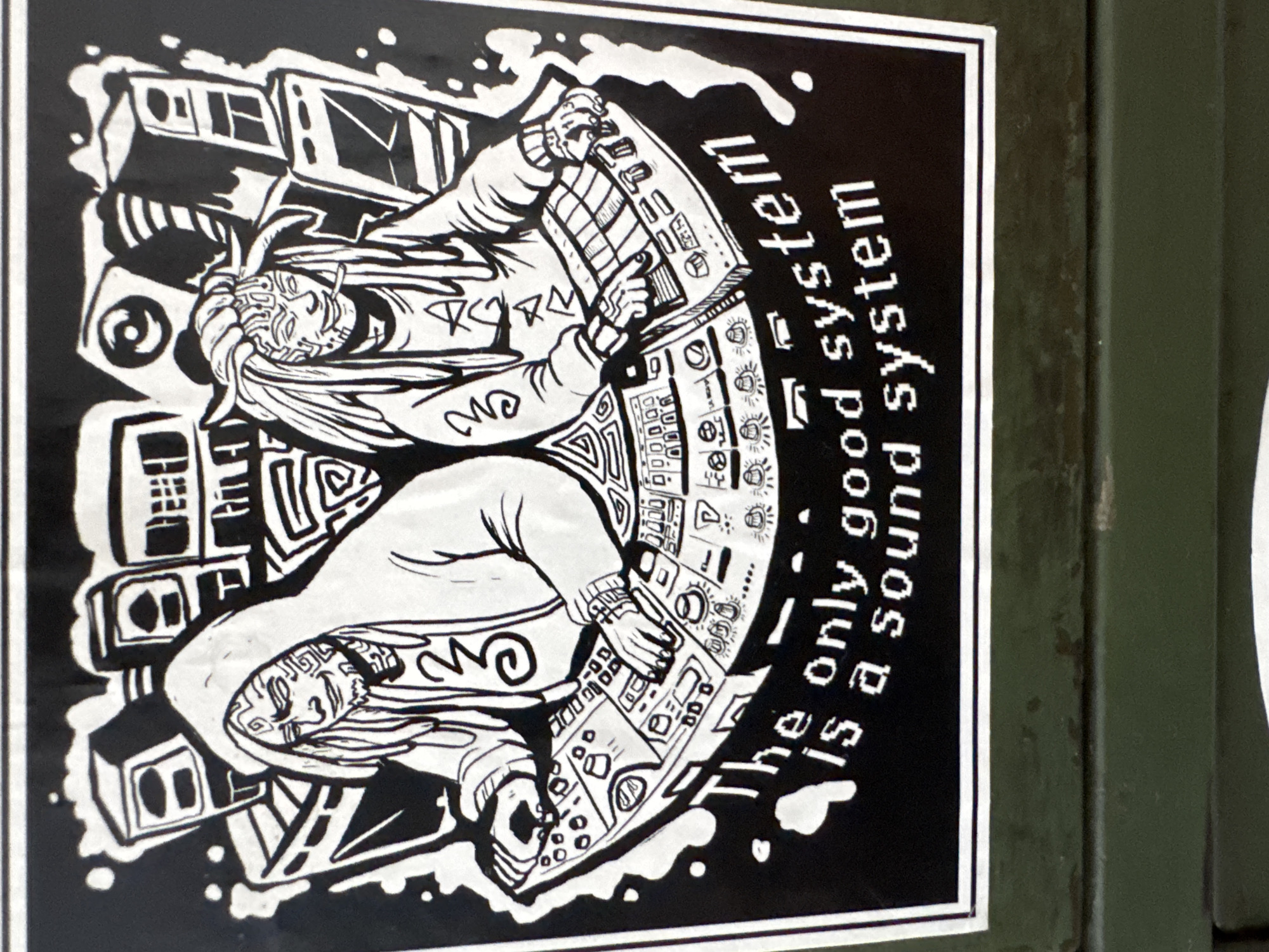 A black and white sticker affixed to a green metal surface in an urban location shows two figures operating a sound system. The figures are stylized with patterned clothing and facial features. The sound system is detailed with various knobs, buttons, and lights. The sticker reads, "The only good system is a sound system." The location is Unknown, Unknown.
