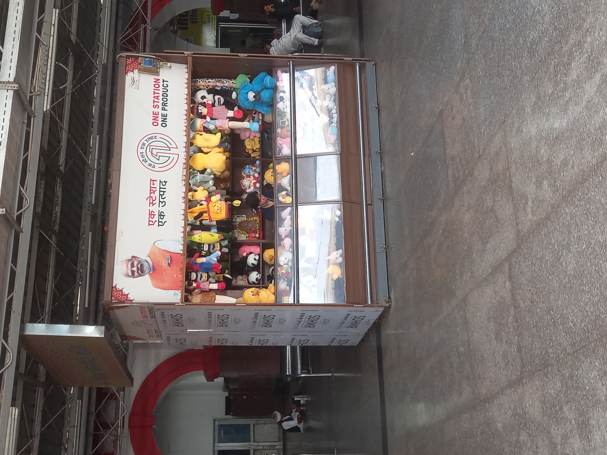 The image shows a "One Station One Product" kiosk at a train station in Jhansi, India. The kiosk is filled with stuffed animals and has an image of Narendra Modi on the side. There are people sleeping on the platform in the background.