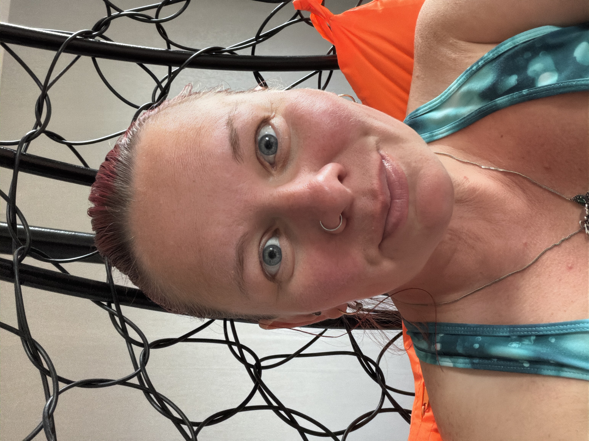 A selfie of a person with blue eyes and light skin, wearing a teal and white top and a silver necklace, with a nose ring, taken in what appears to be a black wire hanging chair, backed by an orange fabric. The location is unknown.