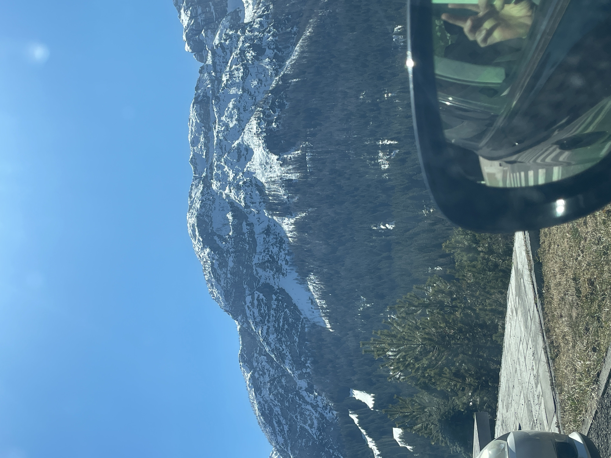 The image shows a landscape in the Sappada/Plodn/Sapade, Italy area, taken from inside a vehicle. A mountain range covered in snow and trees occupies the left and center of the frame, with a clear blue sky overhead. The right side of the image shows part of the vehicle's side mirror reflecting a hand and a portion of the interior. The foreground includes a roadside area with grass and a concrete barrier.