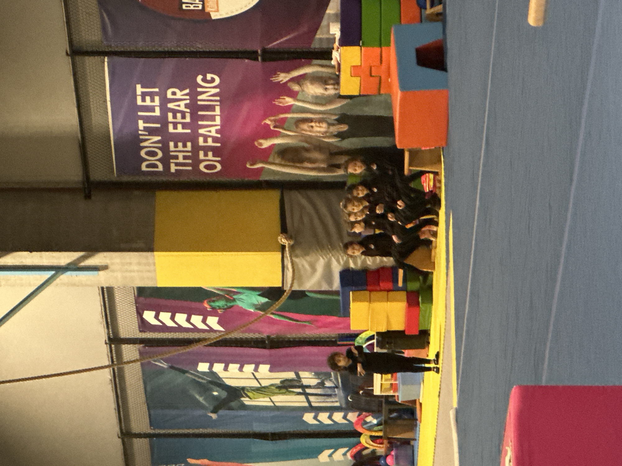 The image shows an indoor gymnastics facility, possibly in Esenyurt, Turkey (though this cannot be confirmed from the image alone). A group of children dressed in black are seated, facing the camera, with their coach standing to the side. Behind them are large, colorful foam blocks used for training, along with safety mats. Banners on the wall display motivational slogans related to gymnastics, such as "Don't let the fear of falling" and feature images of gymnasts. A rope hangs from the ceiling, indicating climbing activities. The facility is brightly lit with an apparent focus on safety and training.