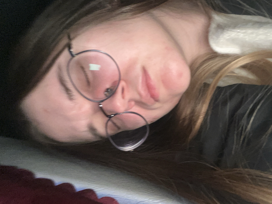 A close-up image depicts one individual, a young adult female with long brown hair, appearing to be asleep or resting, with eyes closed and lips slightly parted. She wears round, wire-rimmed eyeglasses, which reflect a rectangular light source on the right lens. Her skin is fair, with minor blemishes visible on her forehead and chin. She is dressed in a dark-colored garment, possibly a jacket, over a lighter-colored garment visible at the lower right. The blurred background features a vertical, red, wavy object on the left, adjacent to a lighter, possibly off-white, vertical surface. The setting is an indeterminate indoor environment. The scene is located in Dubrava, Russia.
