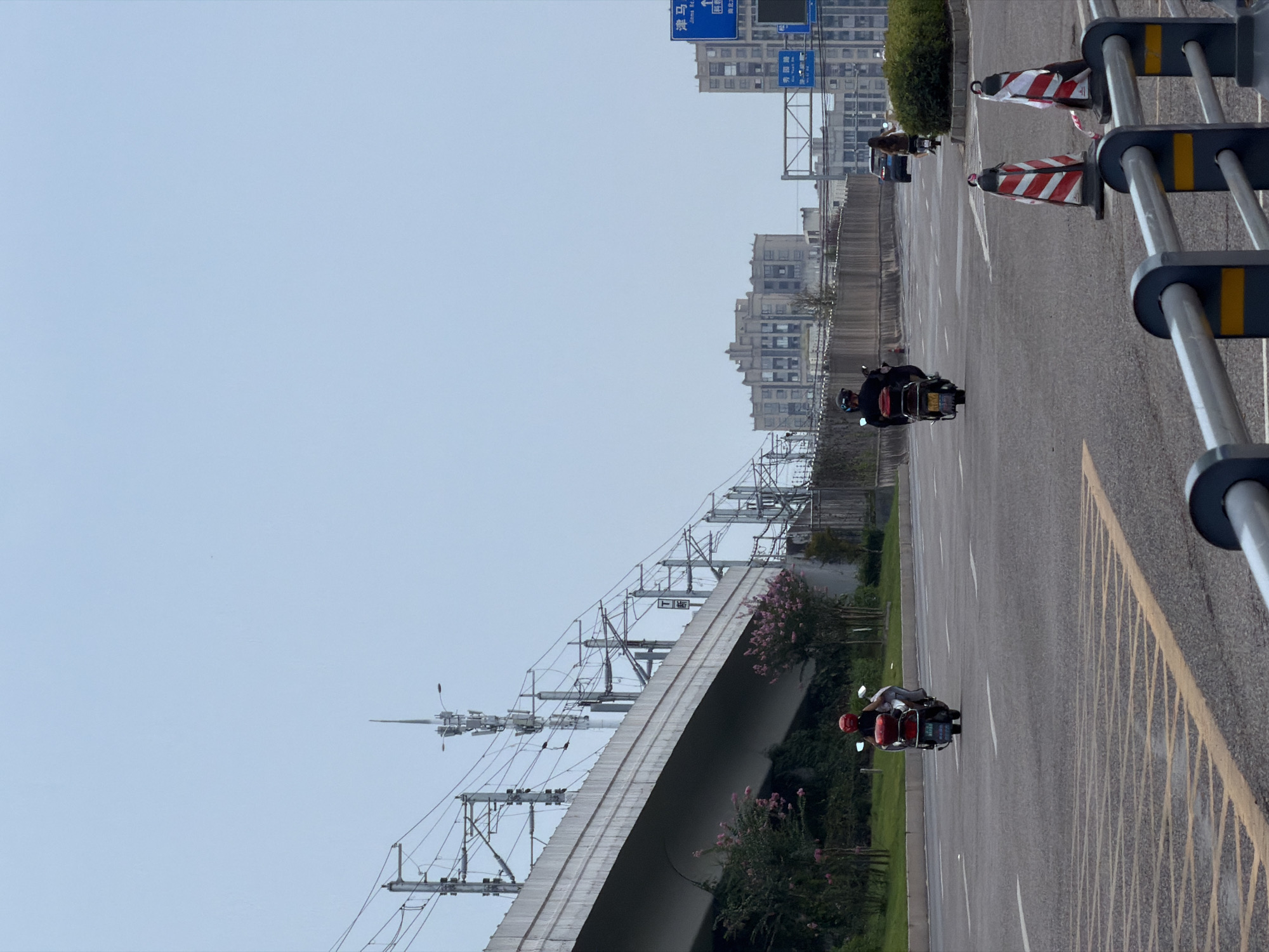 The image shows a multi-lane road in Jiangjin District, Chongqing, China, under an overcast sky. A concrete overpass, likely for a railway or elevated road, dominates the left side of the frame, supported by visible pylons and adorned with numerous overhead power lines and a tall communication tower. Pink flowering bushes are visible beneath the overpass.
On the road, several individuals are riding motorcycles. In the foreground on the left, a person on a red scooter with a white backrest is moving away from the camera. Further down the road, another individual on a black scooter is also traveling in the same direction. On the right side of the road, a woman on a scooter is seen from behind, and a person on a black scooter is also visible. A dark colored car is driving in the distance. Traffic cones are placed on the right side of the road in the foreground.
In the background, several multi-story buildings, including a large, distinctively shaped building, are visible. Blue traffic signs with Chinese characters are present, indicating street names or directions. The road surface is asphalt with clear lane markings. A metal barrier with reflective yellow and grey stripes runs along the right side of the foreground.