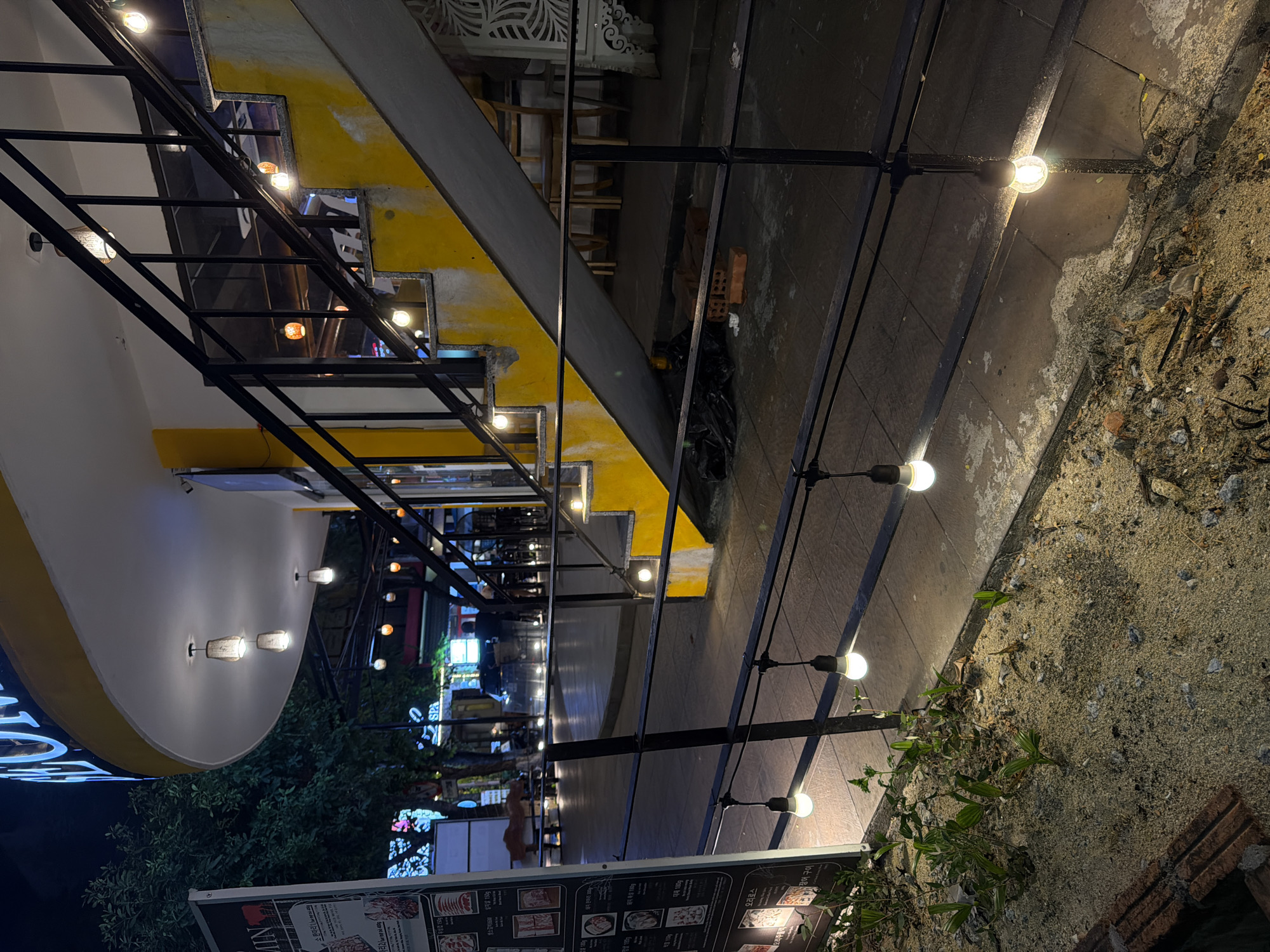 Here is a detailed description of the image:
**Scene:** The image presents an outdoor restaurant or dining area at night, likely located in a bustling urban area. String lights are strung across the space, casting a warm glow.
**Subjects & Objects:**
* **Building Architecture:** A structure is visible, featuring a yellow and gray color scheme, with a staircase element.
* **Lighting:** Numerous string lights are strung across the space, along with fixed light fixtures on the building facade.
* **Menu Board:** A menu board is situated at the bottom, displaying food items and pricing. The menu includes Korean text, but also words in English, such as "steak".
* **Vegetation:** Green foliage and plants are visible in the lower left, adding a touch of nature.
* **Street/Sidewalk:** Paved surface and an area of what appears to be dirt, gravel, and debris are present on the right.
**Actions & Interactions:**
* Given the menu board and lighting, the scene is set for dining and relaxation.
**Environment:**
* The image is taken at night, with the lighting creating an intimate atmosphere.
* The location is most likely an urban street, considering the architecture, paving, and the presence of restaurants.
**Location:**
The presence of Korean text on the menu, along with English and Vietnamese, suggests the image might be taken in a location with an international community or tourist area. Given the Vietnamese on the menu, it may be possible this restaurant is located in the city of Đà Nẵng, Vietnam.