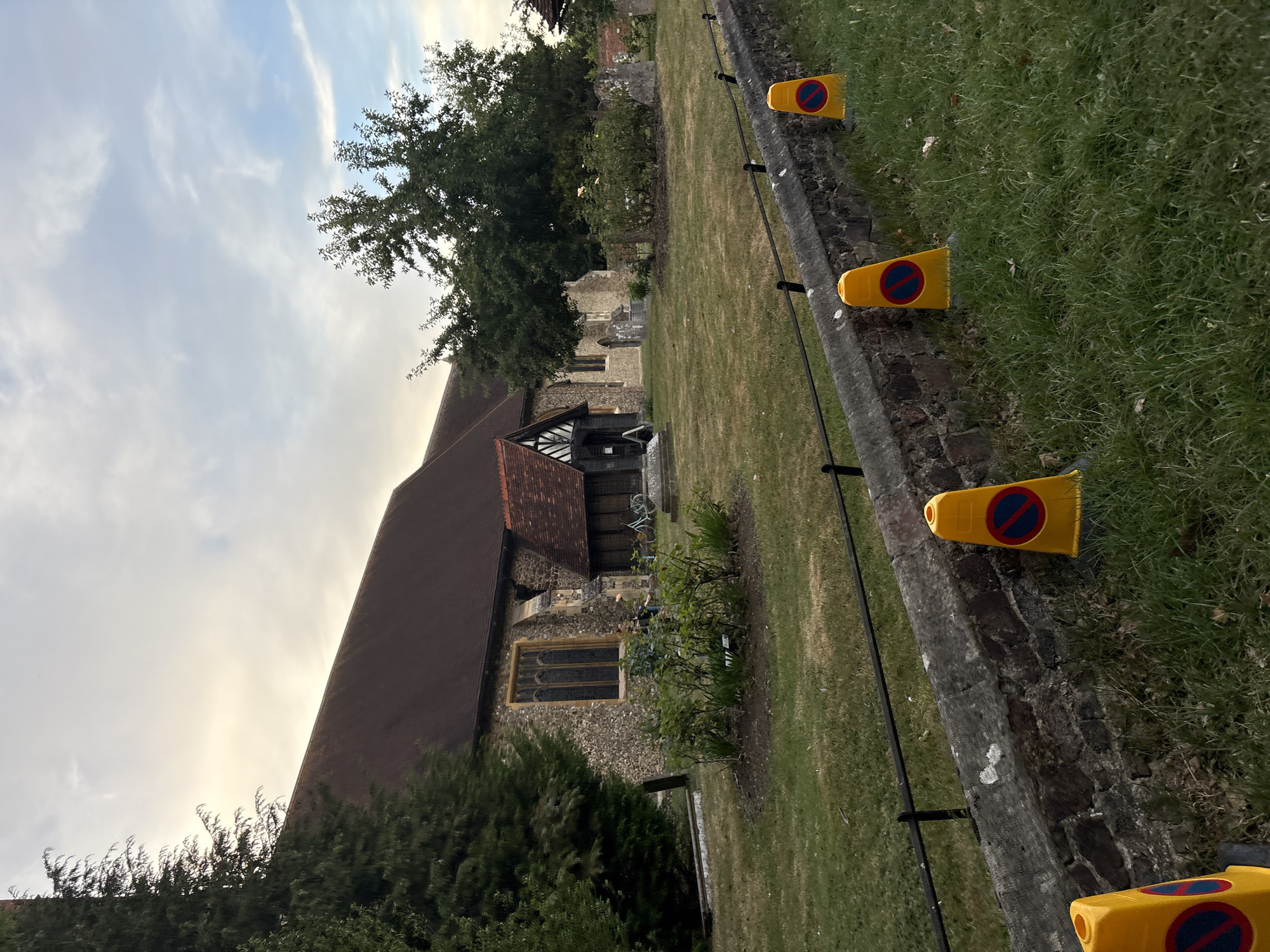 A view of St. Mary's Church, Chelmsford, United Kingdom, taken from the roadside. The church has a prominent stone facade with a pitched, dark tile roof. In the foreground, a low stone wall separates the grassy churchyard from the road. Several yellow traffic cones with a red and blue prohibition symbol are placed along the edge of the wall. Trees and gravestones are visible in the churchyard. The sky above is overcast with patches of light.