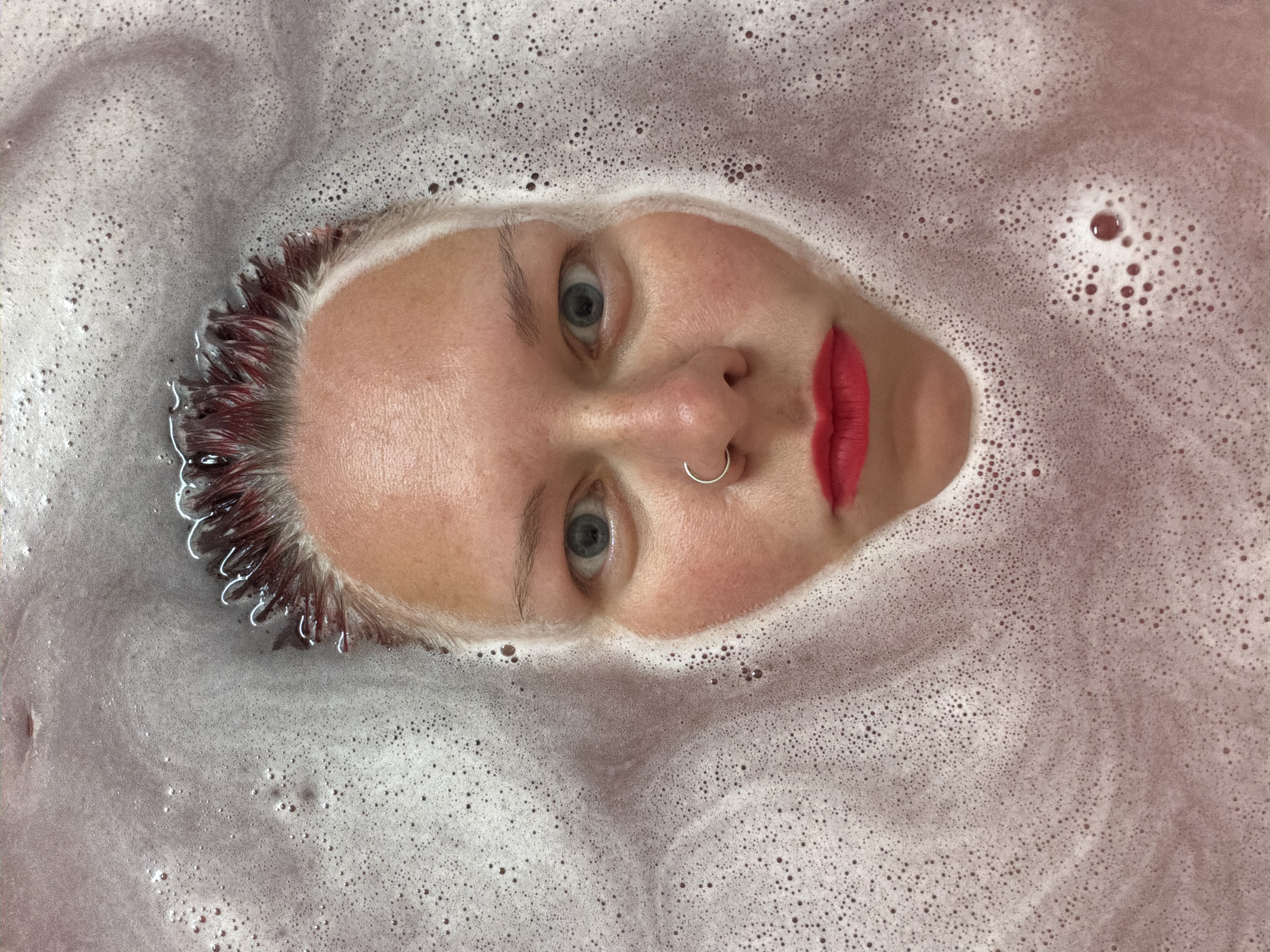 A woman's face is surrounded by water and bubbles in a close-up shot. She has fair skin, blue eyes, and red lipstick. A silver nose ring is visible. Her hair appears wet and dark at the roots, possibly dyed. The water around her face is bubbly and tinted with a reddish-purple hue. The bubbles are small and densely packed. No specific details indicate the location is Chaikovsky, Russia.