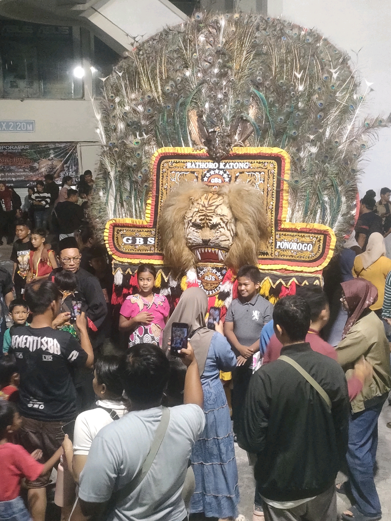 This is a medium shot photograph of a crowd gathered around a large, ornate float decorated with peacock feathers and a lion's head. The float appears to be a cultural or festival display, with the words "BATHORO KATONG" and "PONOROGO" visible on it. A sign in the background reads "MAX. 2.20M" and "OPORAPAR SURABAYA," indicating the location as Surabaya, Indonesia.
In the foreground, several people are visible, many of whom are holding up their phones to take pictures or record videos of the float and the surrounding activity. A young boy in a gray polo shirt with a lion emblem smiles and looks towards the float. A woman in a pink dress stands to his right, her hand on her chest. Behind them, a man in a black shirt and glasses is speaking to someone.
The crowd is diverse, with people of various ages and attire. The environment appears to be indoors or under a covered structure, with artificial lighting contributing to the scene. The overall atmosphere suggests a lively public event.