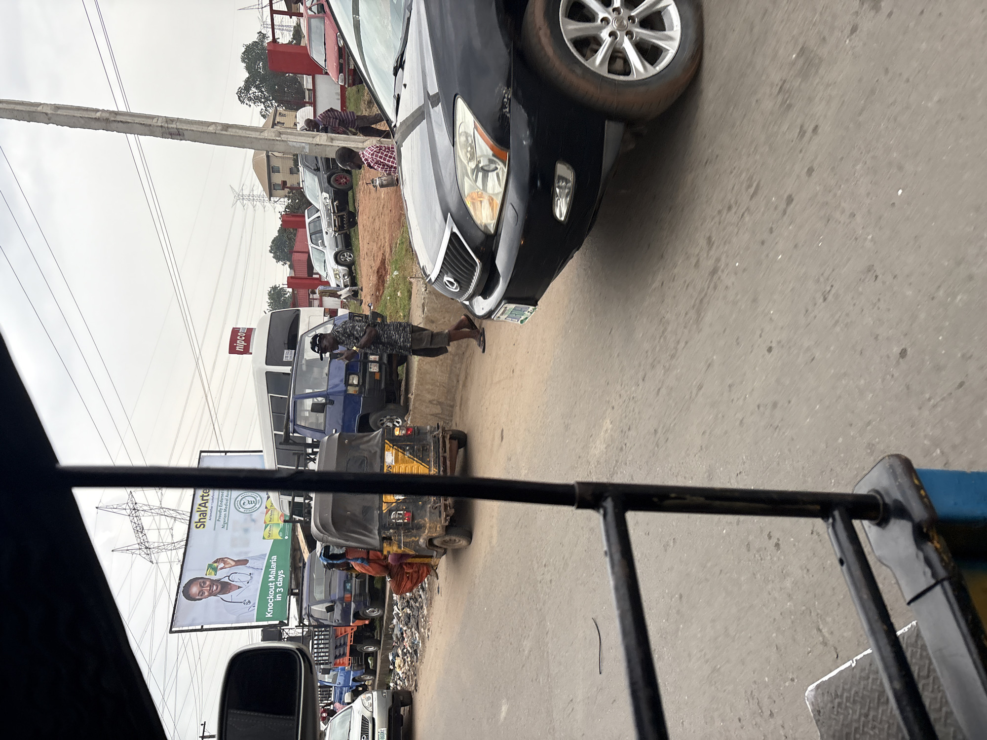 The image depicts a street scene in Onitsha, Nigeria, likely near the Niger Bridge approach, characterized by traffic congestion and urban infrastructure. In the foreground, a black Lexus SUV is prominently positioned, its front right wheel and headlight visible. The vehicle appears to be in motion or stopped in traffic. Behind the SUV, a yellow and black auto-rickshaw is visible, with a person in orange clothing seated inside.
Further down the road, a variety of commercial vehicles are present, including blue trucks and a white bus. A large billboard advertising "Shal'Arte" with the slogan "Knockout Malaria in 3 days" is a significant element in the background. The billboard features an image of a woman in a doctor's coat holding a product. Adjacent to the billboard, a red sign with "nipco" is visible, suggesting a fuel station or related business.
The street itself is paved and appears to be a busy thoroughfare. Piles of refuse are noticeable along the roadside, indicating urban waste management challenges. Several individuals are seen walking or standing near the vehicles. The overall environment suggests a typical bustling commercial area in a Nigerian city, with power lines and transmission towers visible in the sky, indicating an urban setting with ongoing electrical infrastructure. The weather appears overcast, contributing to a diffused light across the scene.