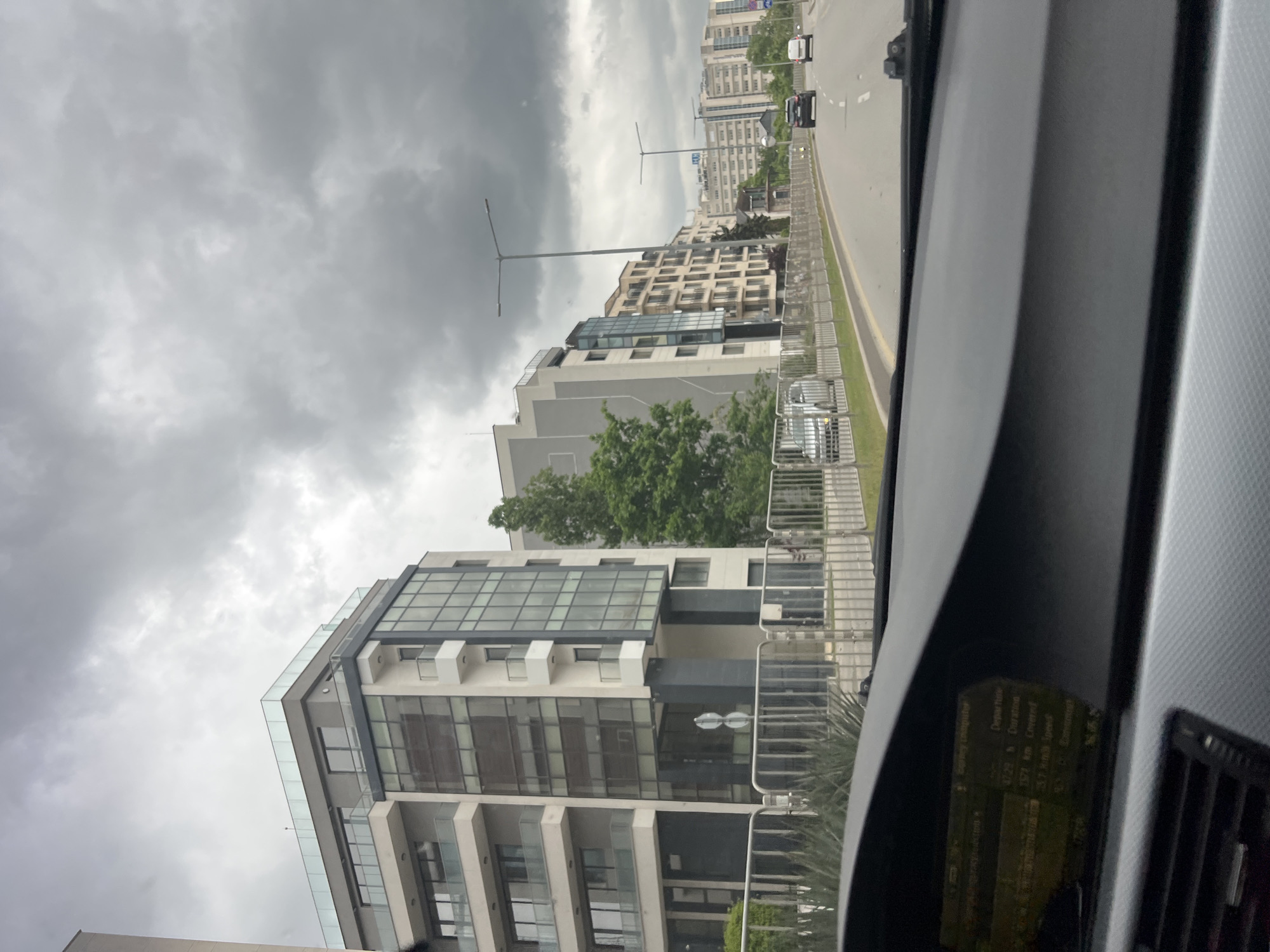 The image shows a view from inside a car, likely from the passenger seat. The car is traveling on a road alongside buildings, seemingly in an urban environment. The sky is overcast with gray clouds. The buildings are modern, multi-story structures with glass and concrete facades. There is some greenery visible, suggesting trees or landscaping. The location is in Sofia, Bulgaria. There are other vehicles on the road.