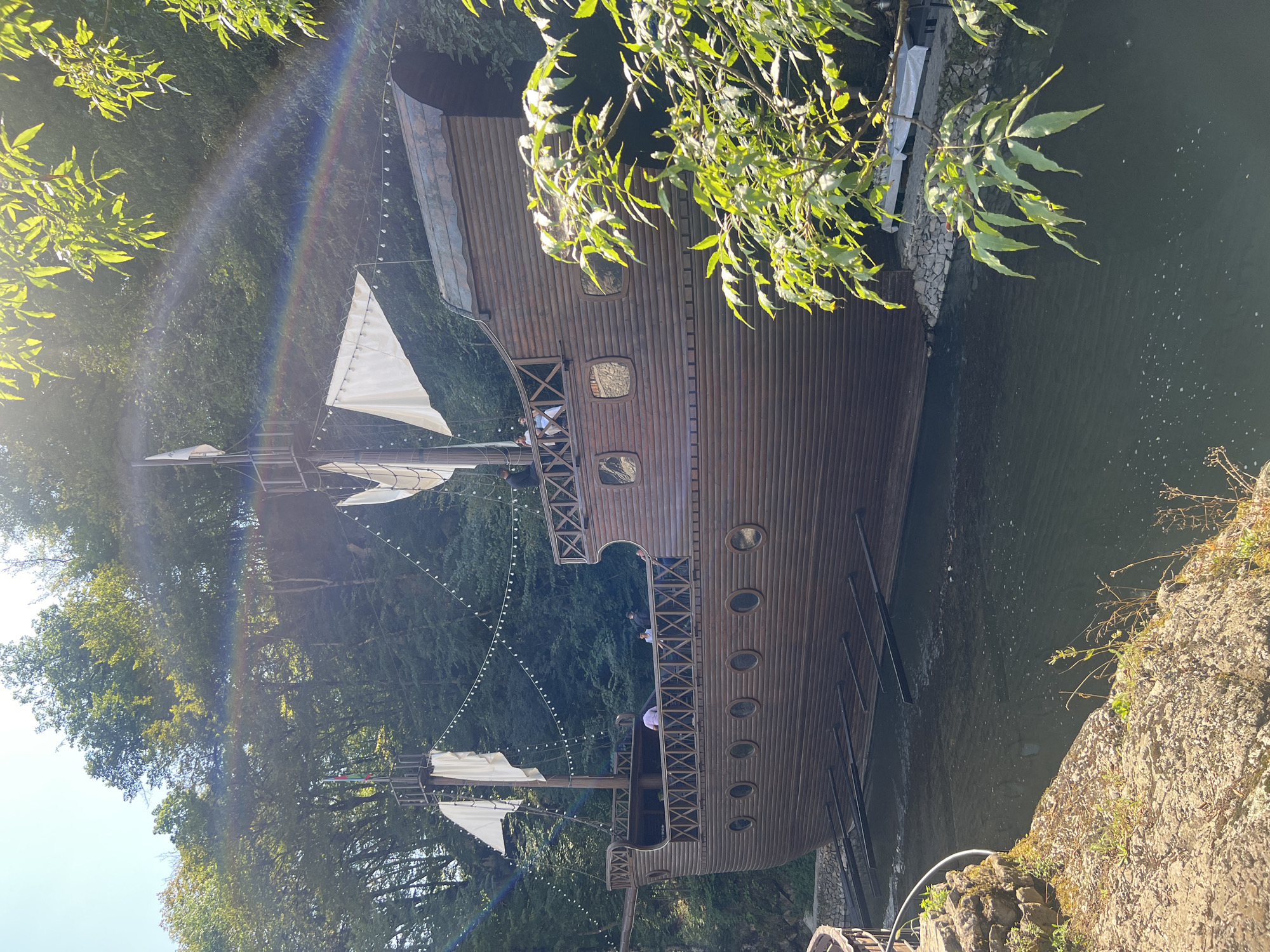 The image depicts a wooden replica of a pirate ship docked in a body of water, surrounded by lush green trees and foliage. The ship is detailed, with several decks, masts, and white sails. String lights are strung along the railings of the ship, suggesting it might be a restaurant or entertainment venue. There are a few individuals visible on the deck of the ship, appearing to be casually observing the surroundings. The water is dark and reflects the trees. In the foreground, a rocky embankment with some vegetation leads down to the water. Sunlight filters through the trees, creating a dappled effect and a faint rainbow visible in the upper right quadrant of the image. Based on the available information, this scene is located in the city of Sarısa, Azerbaijan.