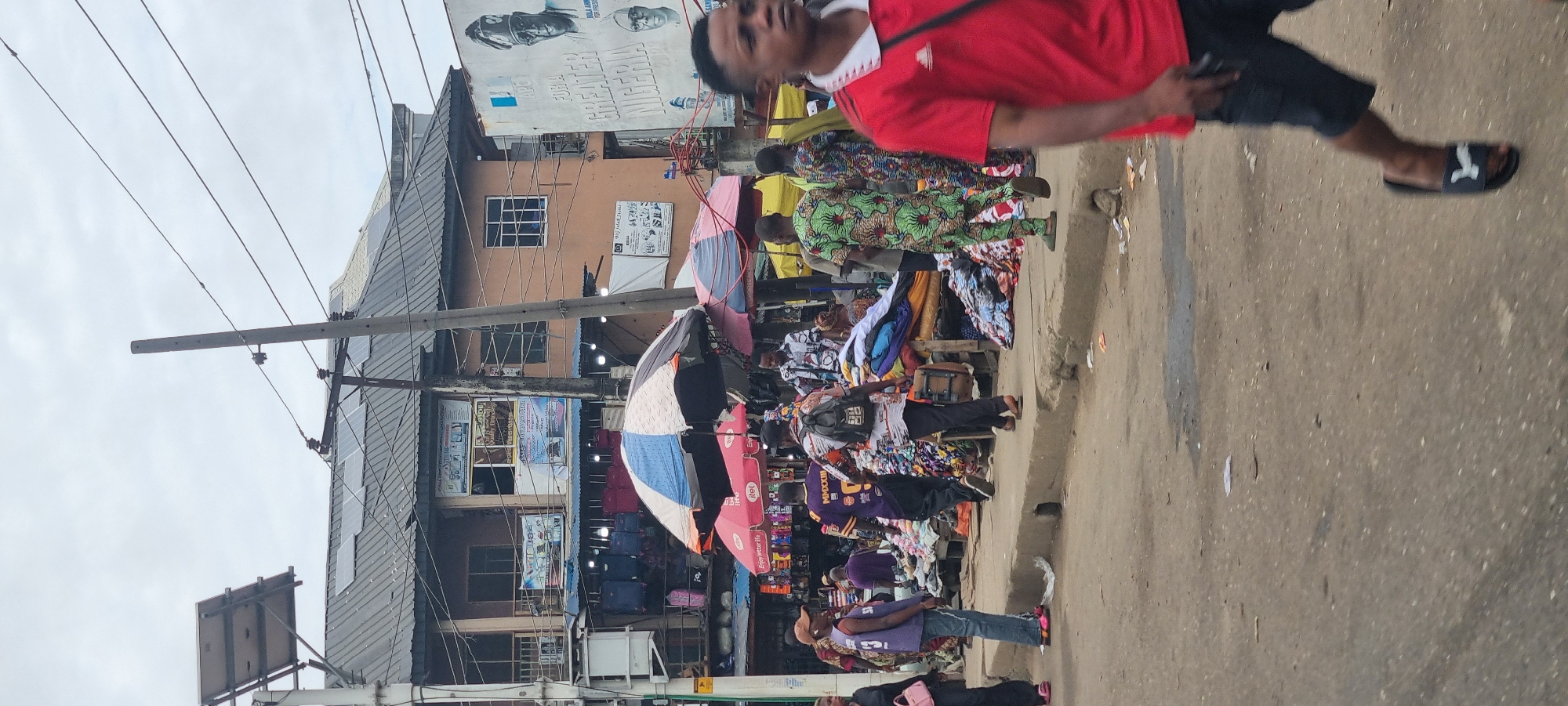 The image captures a bustling street scene in Ikeja, Nigeria, likely a market area. In the foreground, a person wearing a red polo shirt and dark shorts walks away from the camera, with their legs and feet visible. Behind them, a vibrant market is in full swing. Numerous stalls are set up, displaying a variety of goods, prominently featuring textiles, clothing, and various packaged items. Colorful umbrellas and awnings provide shade over the stalls.
People are seen browsing, interacting, and moving through the market. One individual in the middle ground wears a patterned green and white outfit, while another wears a purple t-shirt with "MMXXIII" on the back. A person in a patterned shirt and black backpack is also visible. Further back, a building with multiple floors stands, featuring balconies and windows, with solar panels visible on the roof. Signs and advertisements are posted on the building. The street is paved, with some debris and scattered papers on the ground, indicative of a busy public space. Overhead, utility wires are strung between poles, suggesting the presence of electricity infrastructure. The sky is overcast. The overall impression is one of lively commerce and daily life in an urban Nigerian setting.