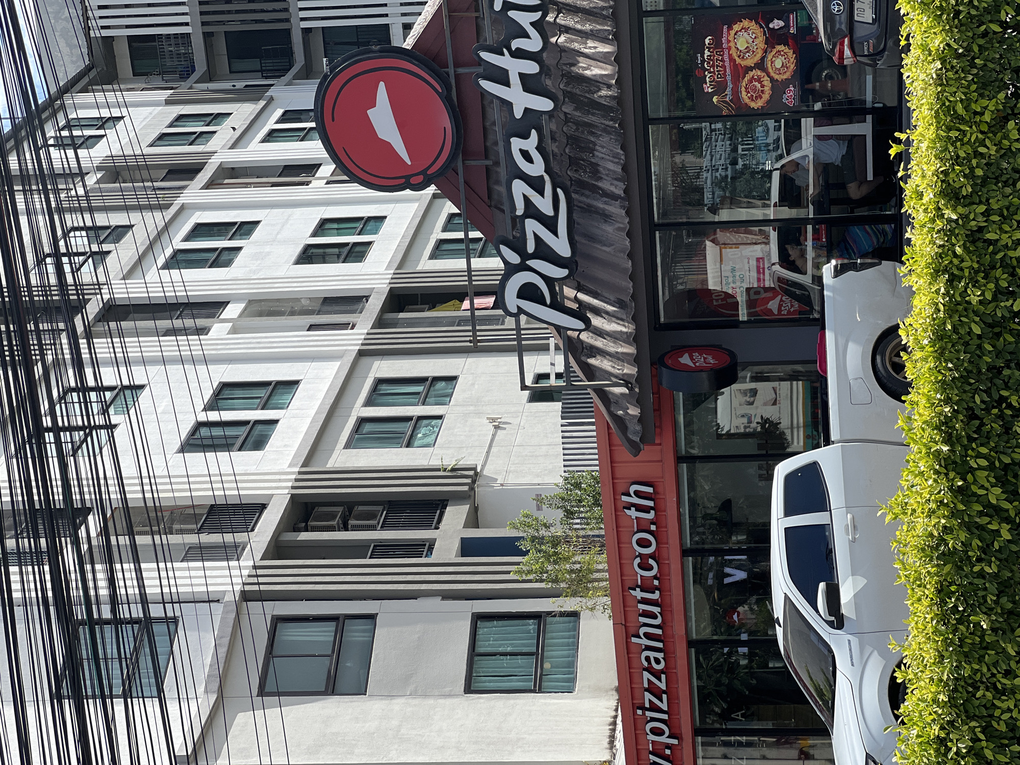 A wide shot captures a Pizza Hut restaurant in a city, featuring a white pickup truck parked in front and a modern apartment building in the background. The Pizza Hut's facade is adorned with its iconic red and white logo and the "Pizza Hut" name in cursive. The restaurant's windows display advertisements for "Volcano Pizza" and show a seating area with two people. The building behind the restaurant exhibits a contemporary architectural style with multiple floors, balconies, and numerous windows. Overhead, a complex network of electrical wires crisscrosses the sky, adding to the urban landscape. A neatly trimmed green hedge runs along the foreground, separating the street from the establishment.
