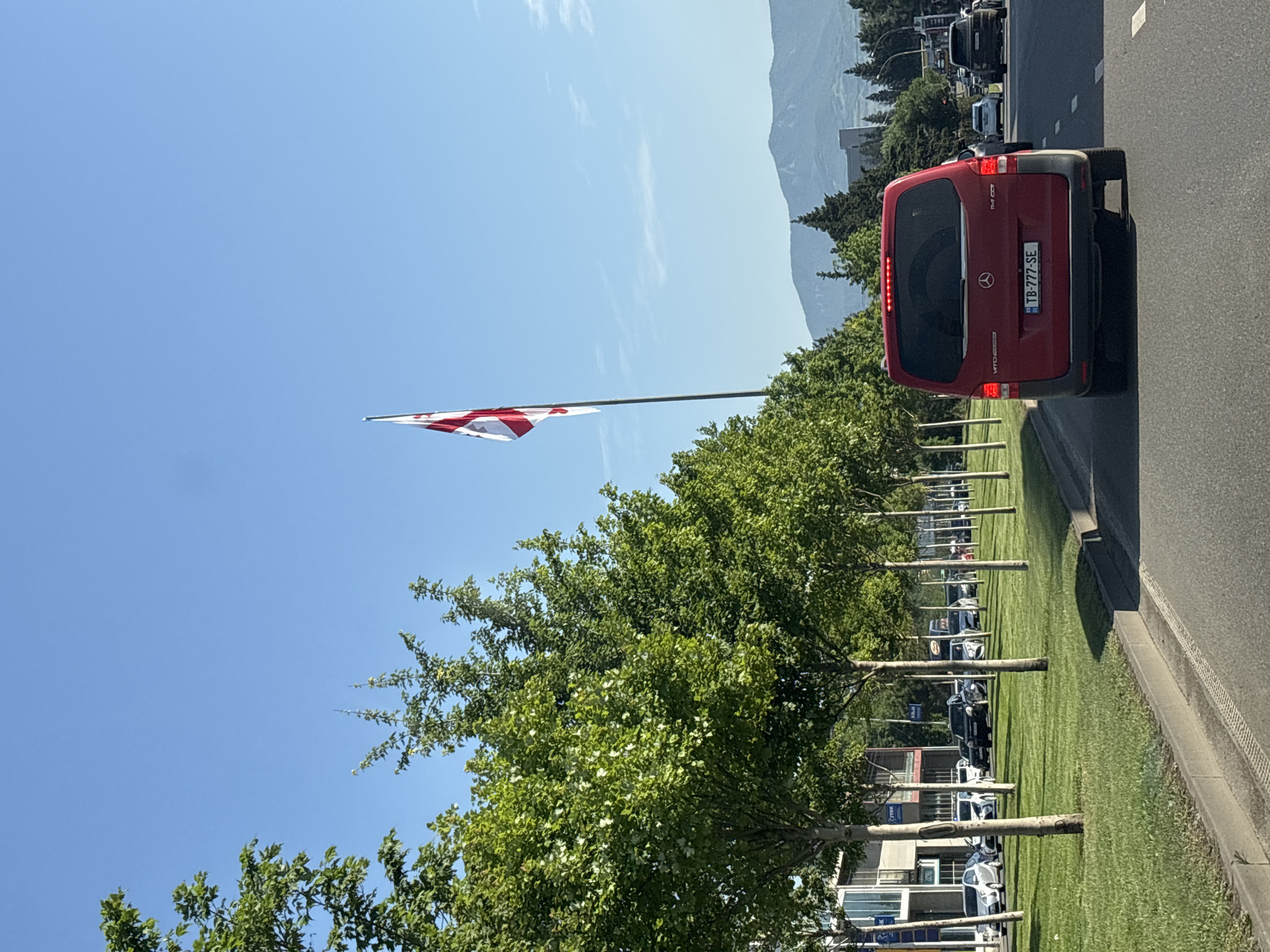 The image shows a street scene in Tbilisi, Georgia, on a clear day. In the foreground, a red van is driving on the road, with its license plate reading "TB-777-SE". To the left of the road, a row of trees lines a grassy median, with modern buildings visible behind them. A Georgian flag is prominently displayed on a flagpole to the right of the trees. In the distance, mountains can be seen. Several other cars are also visible on the road and parked along the side. The scene appears to be an urban or suburban area.