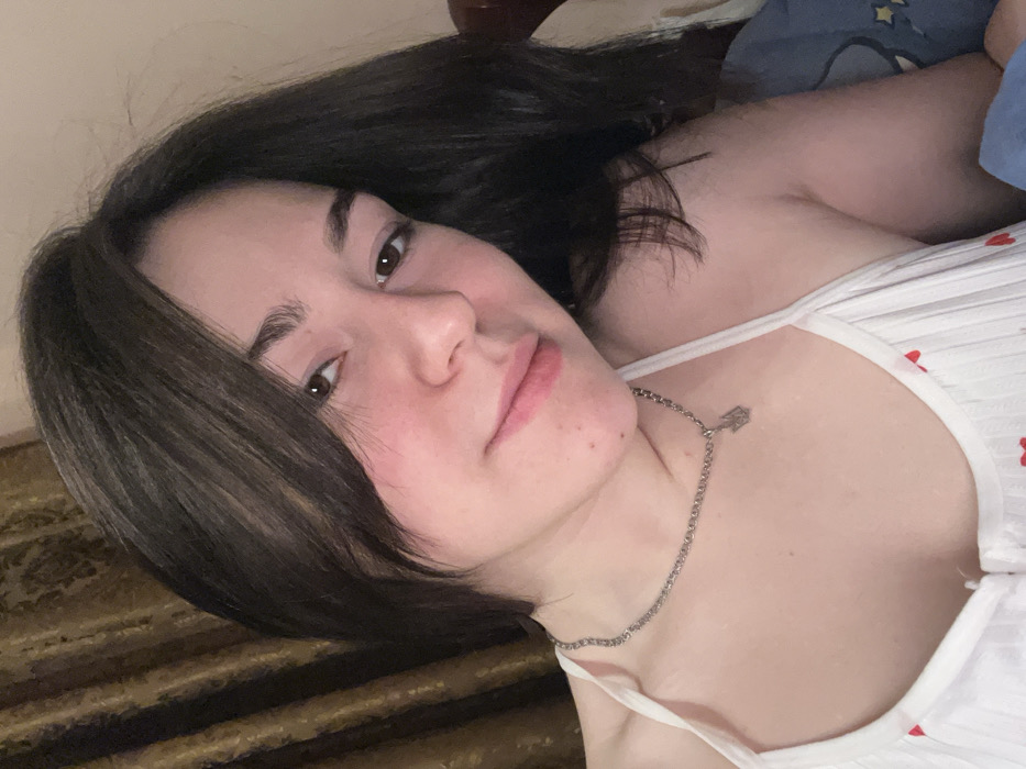 A young woman with dark hair and brown eyes is lying down, gazing directly at the viewer. She's wearing a white tank top with red heart patterns and a silver chain necklace. The background includes a light-colored wall and what appears to be a bed with a blue patterned blanket. The angle suggests she might be taking a selfie.