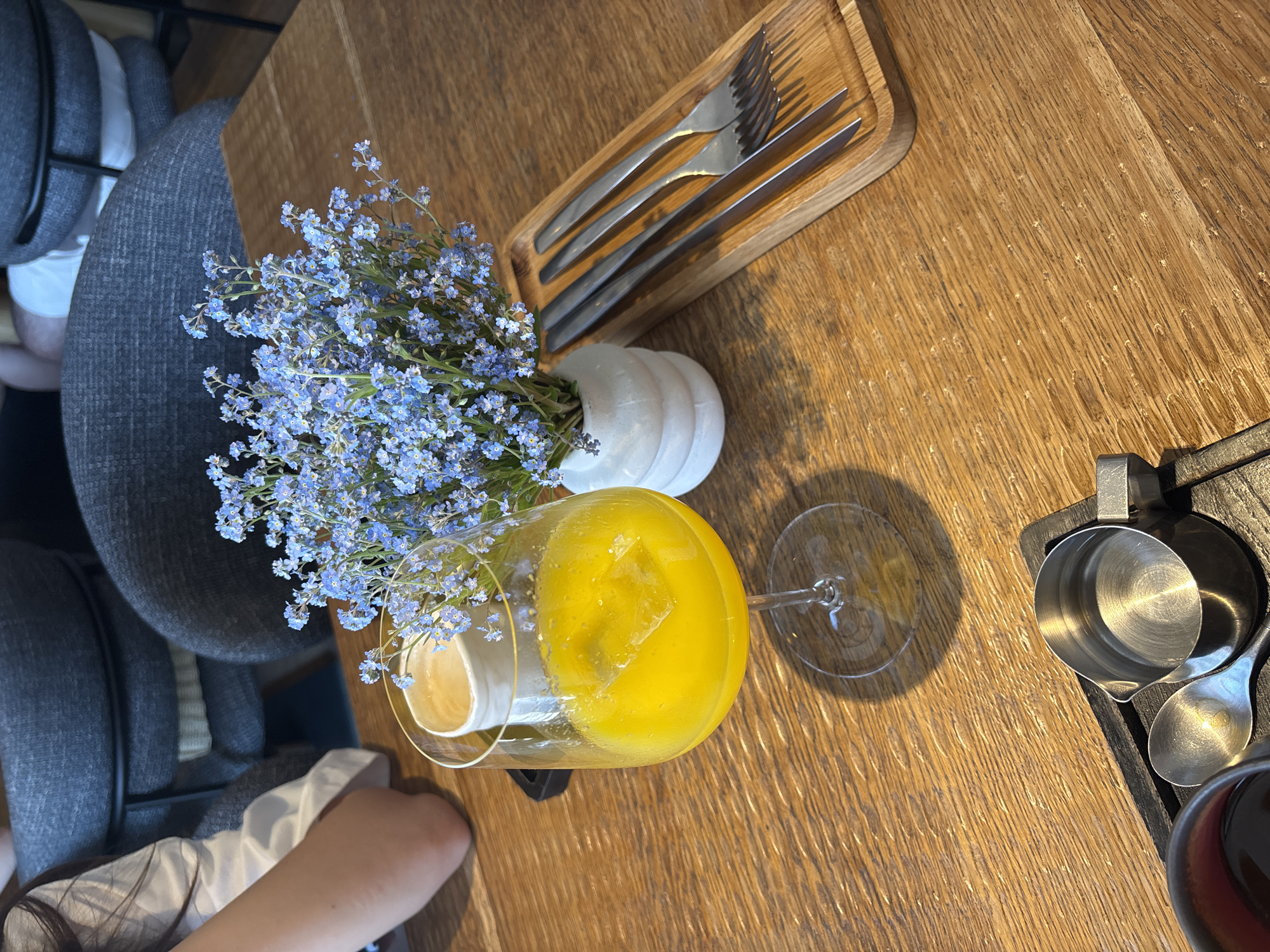 This image captures an indoor scene at a dining establishment in Krasnoyarsk, Russia, featuring a wooden table set with drinks, flowers, and cutlery. The table is made of light brown wood with a prominent grain pattern. In the left foreground, the lower arm and elbow of a person wearing a white shirt are visible, indicating they are seated at the table.
Prominently displayed in the center is a clear, stemmed wine glass filled with a bright yellow liquid, containing at least one large, square ice cube. Behind this glass, slightly to the right, rests a small, white, horizontally ribbed ceramic vase holding a compact bouquet of numerous small blue flowers, consistent with forget-me-nots. Partially obscured behind the yellow drink and near the flowers is a small white cup containing a light brown, frothy liquid, indicative of a coffee beverage.
On the right side of the table, a rectangular light wooden tray holds four pieces of dark metallic cutlery: two knives and two forks, arranged parallel to each other. In the bottom left corner, part of a dark cup with a reddish-brown interior rim is visible. Adjacent to it, on a black tray, is a small metallic pitcher filled with clear liquid and a metallic spoon.
In the blurred background, several individuals are seated in dark grey upholstered chairs. One person is visible wearing a white shirt and possibly light-colored shorts. The overall setting suggests a casual yet refined cafe or restaurant environment.