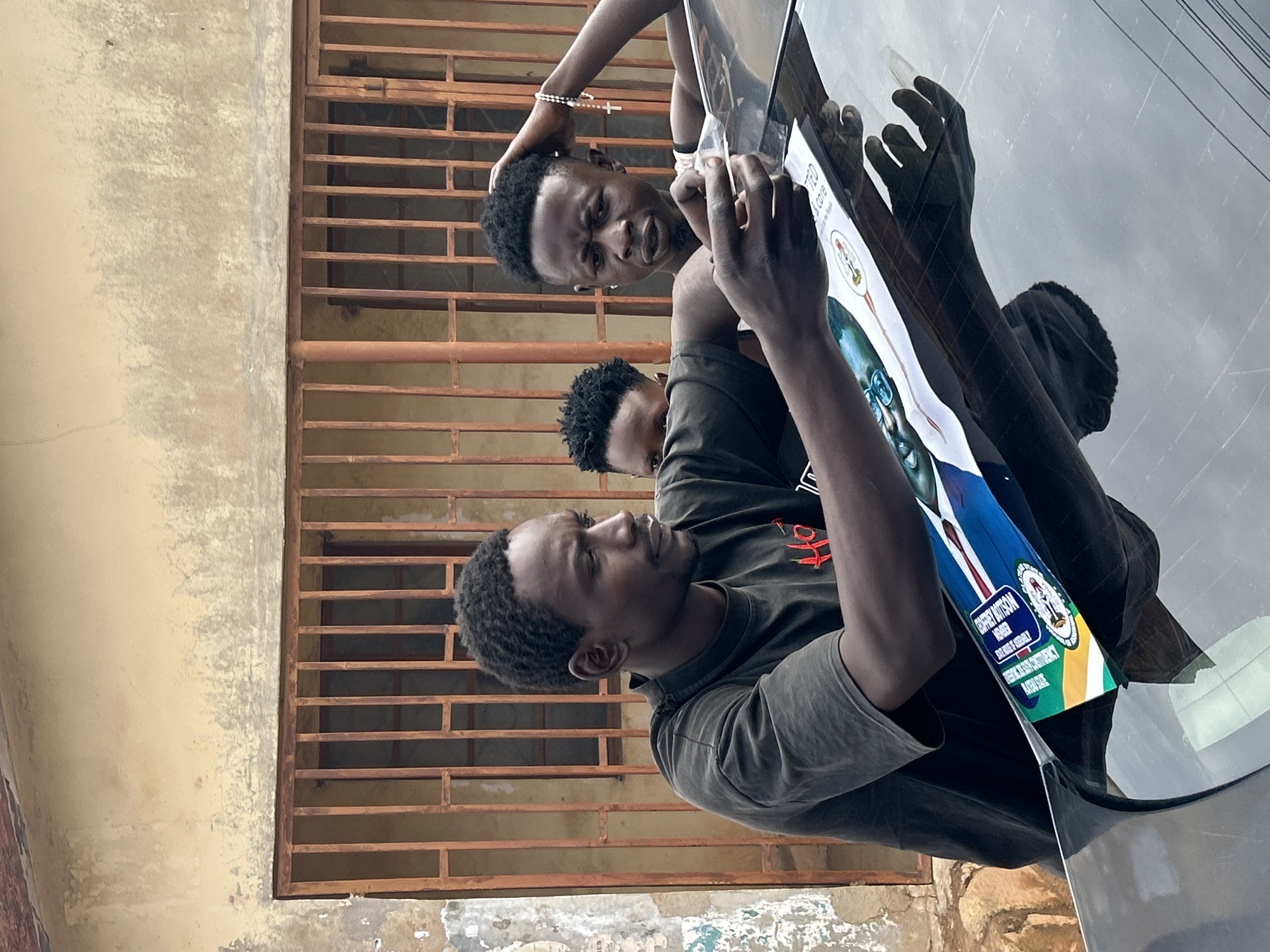 Three Black African young men are standing beside a car, attaching a political campaign poster to the car window. The poster features a man in glasses and a suit. Text on the poster reads "GODFREY BUTRUS M MAGAJI, FOR PLATEAU STATE HOUSE OF ASSEMBLY, PANKSHIN NORTH CONSTITUENCY, PLATEAU STATE". The location appears to be in Jos, Nigeria, judging from the text on the poster. The background shows a weathered building with a metal barred window.