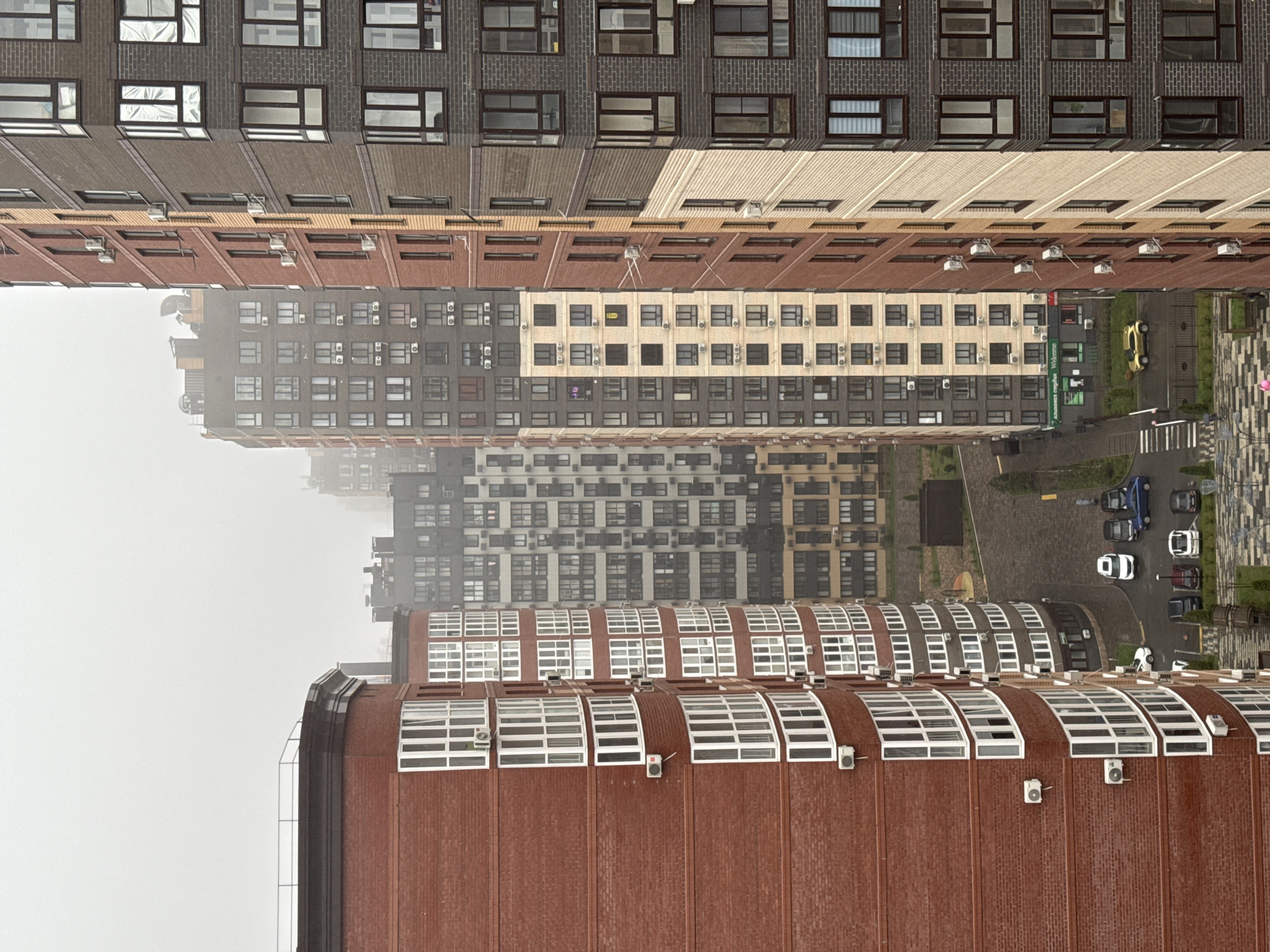 The image shows an urban view between tall buildings in Ovoshchnoy, Russia, likely taken from a high vantage point. The buildings are varied in color and architectural style, constructed of brick and featuring numerous windows, with some equipped with air conditioning units. The lower buildings are visible in detail, with red brick, while the upper buildings vary in tone, from light brown to grey and dark brown, with visible brickwork and similar window patterns. The upper portion of the view is obscured by fog. Below, a courtyard or street with parked cars, trees, and some walkways is visible. A few pedestrians can be discerned. The weather appears to be overcast.