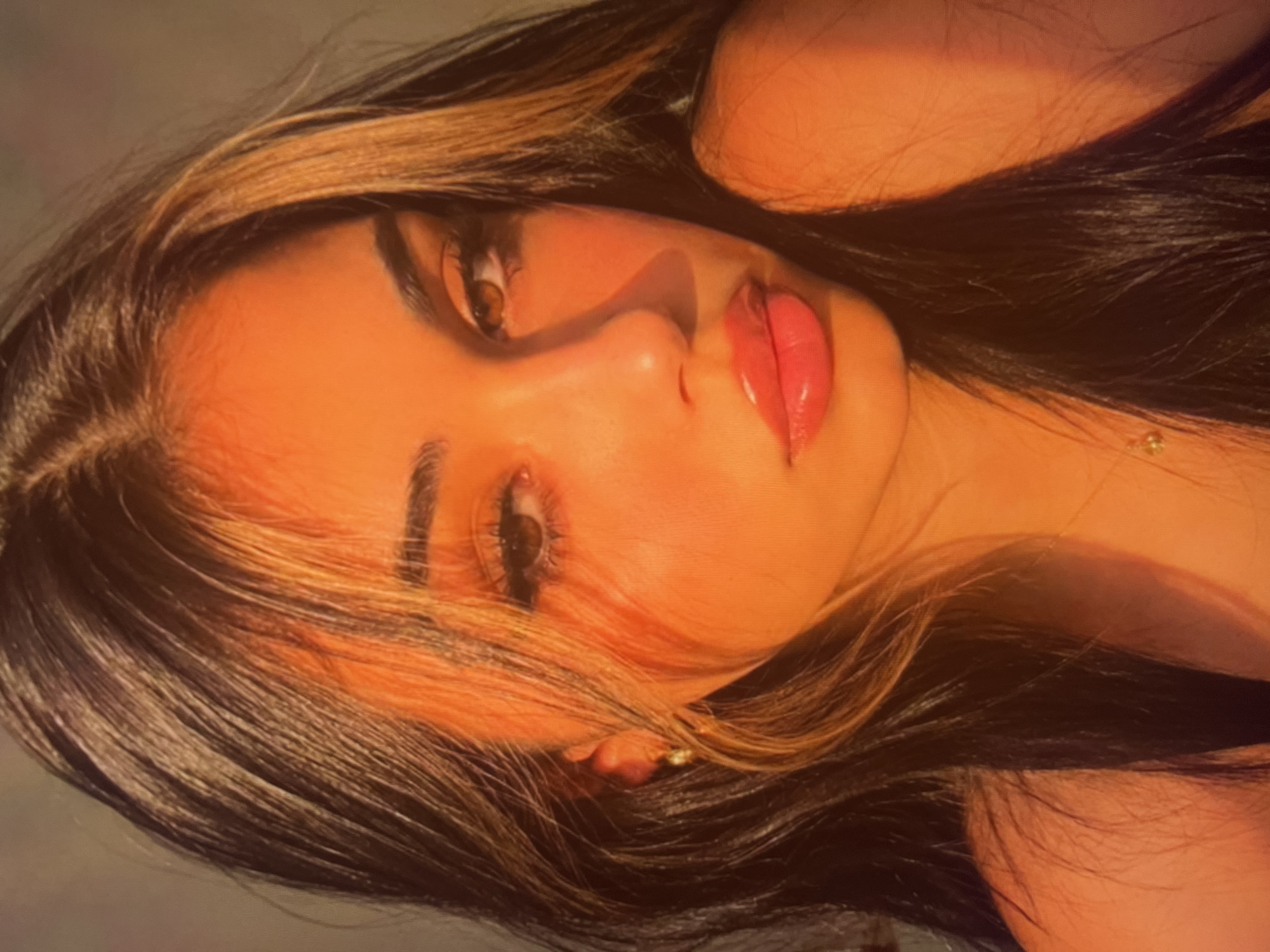 An extreme close-up shot focuses on the face of a young woman with dark hair, illuminated by warm, golden-hour lighting. Her skin is glowing, with a prominent blush and glossy lips. Her eyes, a warm brown, are softly gazing forward, framed by dark, elongated eyelashes and well-defined eyebrows. A subtle gold earring is visible on her left ear. The background is a soft blur of warm tones, with no discernible urban features. The lighting suggests an outdoor setting, possibly during sunset or sunrise. The image is composed with the woman's face as the central subject, creating an intimate and artistic portrait.