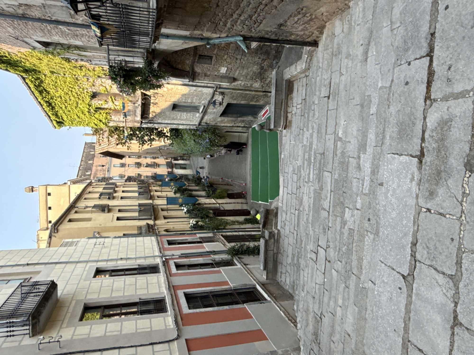 The image captures a downward view of a stone staircase leading into a narrow, European-style street in Girona, Spain. The buildings flanking the staircase are old, with ornate balconies and windows. On the left, a building displays a facade with alternating light and dark horizontal bands, and red and white vertical stripes. On the right, a building is covered in lush green ivy, with a dark wooden door and a lantern above it. The street below features a green surface laid out in parallel strips, possibly a temporary sports court or decorative element. A few orange traffic cones are visible along the street. The sky is clear and blue, suggesting a sunny day. There are no people clearly visible in the immediate foreground, but a distant figure can be seen walking in the street.