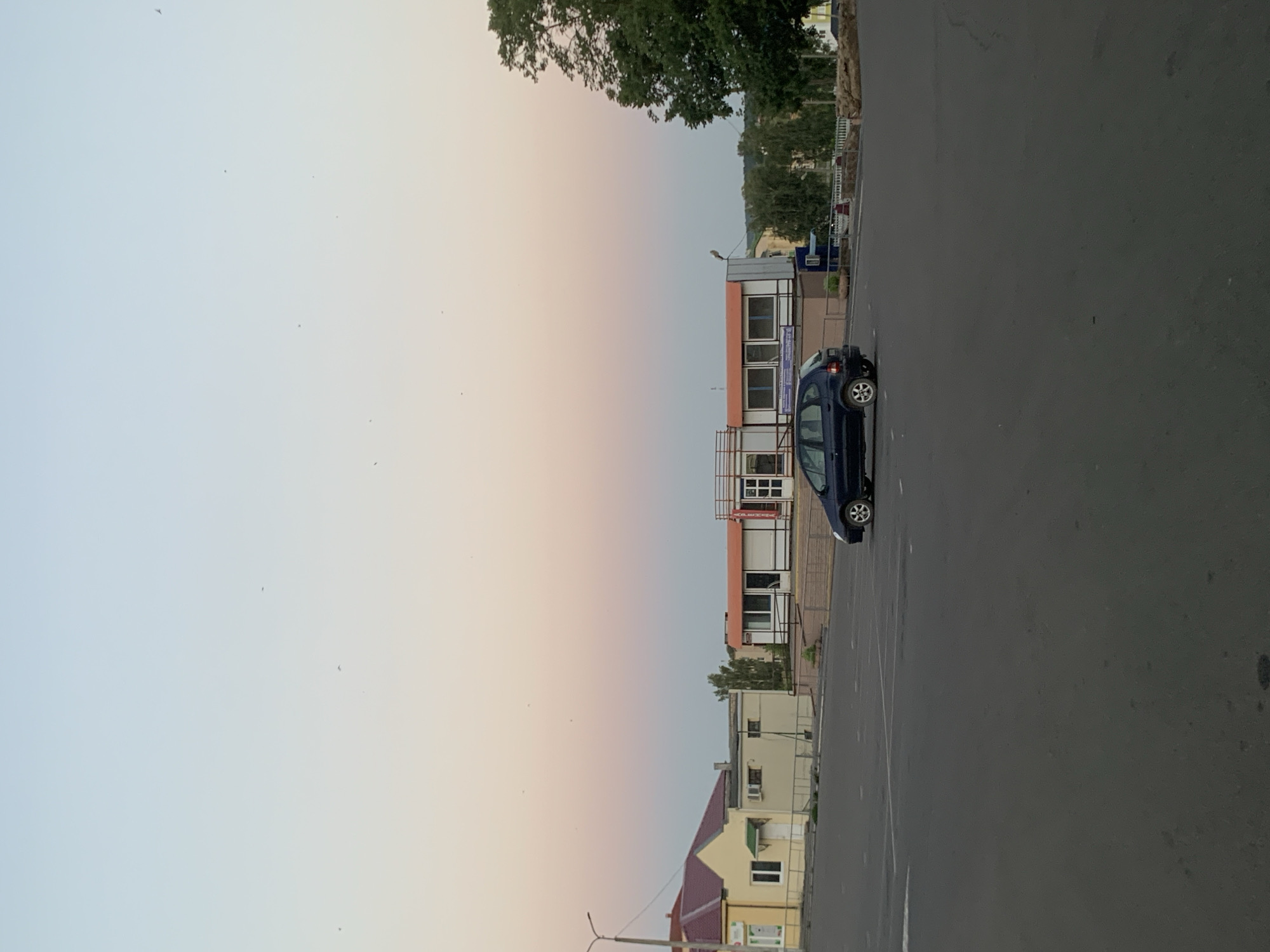 The image captures a street scene at dusk in Kapyĺ, Belarus. The sky displays a gradient of soft blues and pinks, indicative of sunset or sunrise, with several small birds visible against the sky. In the foreground, a dark asphalt parking lot occupies the majority of the frame. A dark blue car is parked on the lot, angled slightly towards the viewer.
In the middle ground, a low-rise building with a white facade and orange trim is situated behind a metal railing. This building appears to be commercial or administrative, with multiple windows and a covered entrance. To the left of this building, a smaller, lighter-colored building with a red roof is visible, featuring a window and a door. Further to the right, behind the main commercial building, a dense green tree stands prominently. The background consists of more trees and hints of other structures, suggesting a suburban or semi-urban environment. The overall impression is one of quietude and the transition of day to night.