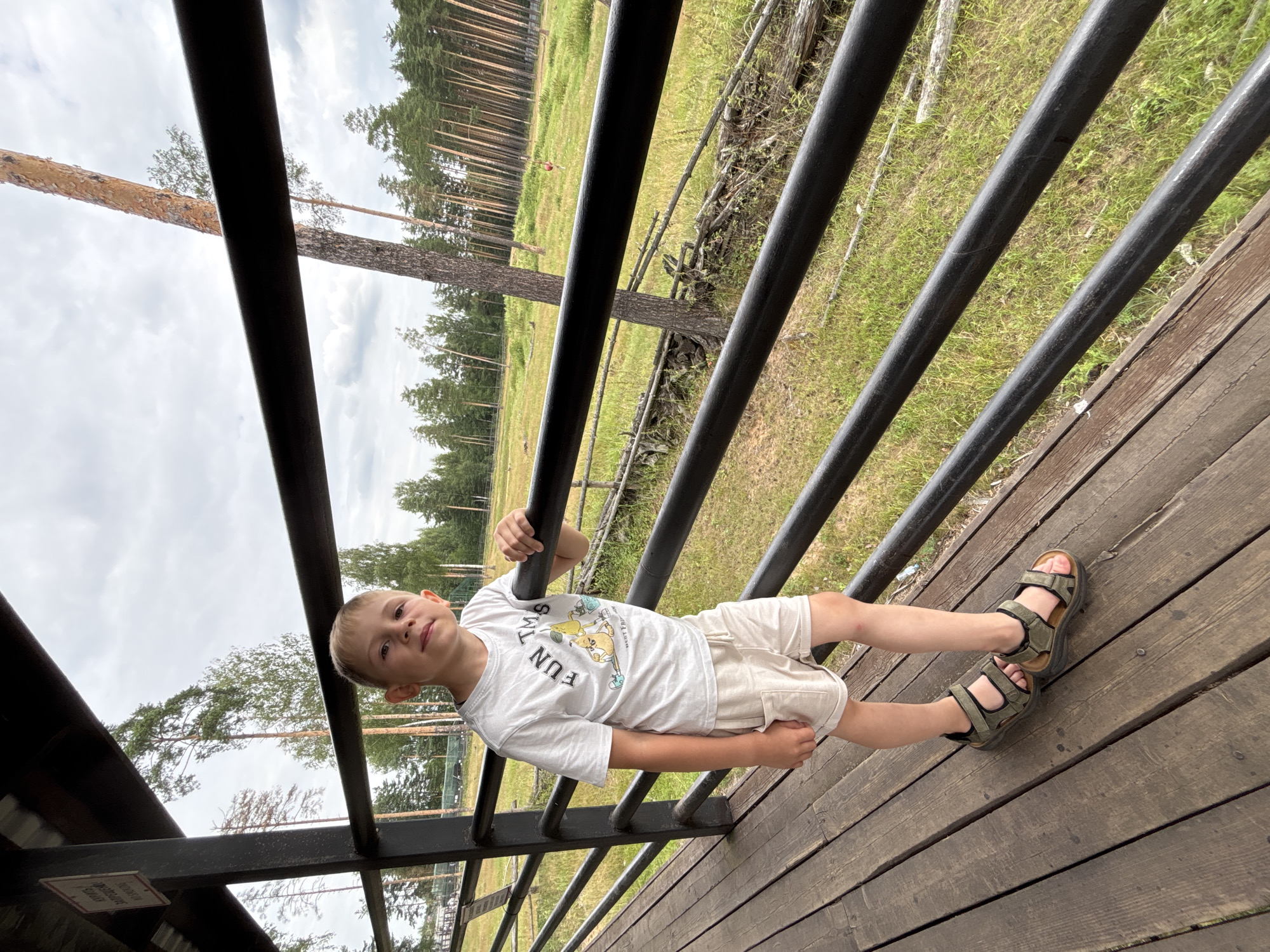 A young boy stands on a wooden deck, leaning against a black metal railing. He is wearing a white t-shirt with a cartoon graphic, beige shorts, and green sandals. He has blond hair and is looking towards the camera with a neutral expression. Behind him, a grassy field with scattered trees and a forest is visible under a cloudy sky. The scene appears to be outdoors, possibly at a park or recreational area in Yaroslavl, Russia.