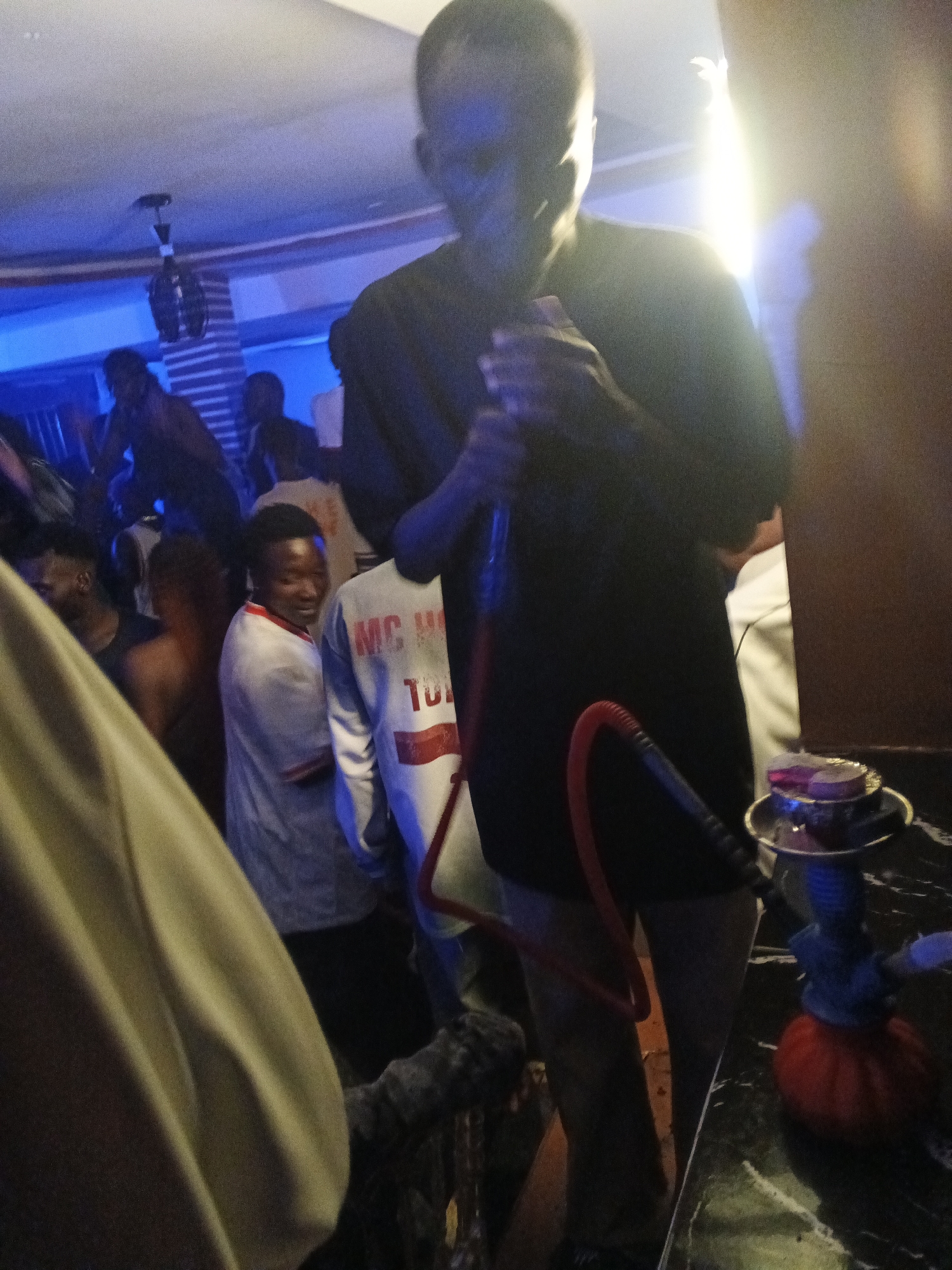 In an indoor establishment, potentially a club or lounge, a group of individuals is gathered. The lighting is dim, with prominent blue and yellow hues illuminating the scene. A man in the foreground, facing away from the camera, holds a shisha pipe with a red hose. The shisha apparatus itself is visible on a dark, reflective surface, complete with glowing charcoal. Other people are present in the background, some of whom appear to be dancing or socializing. One individual in the mid-ground is wearing a white t-shirt with red trim and text that reads "MC HO TO". The environment suggests a lively social setting, possibly a nightlife venue.