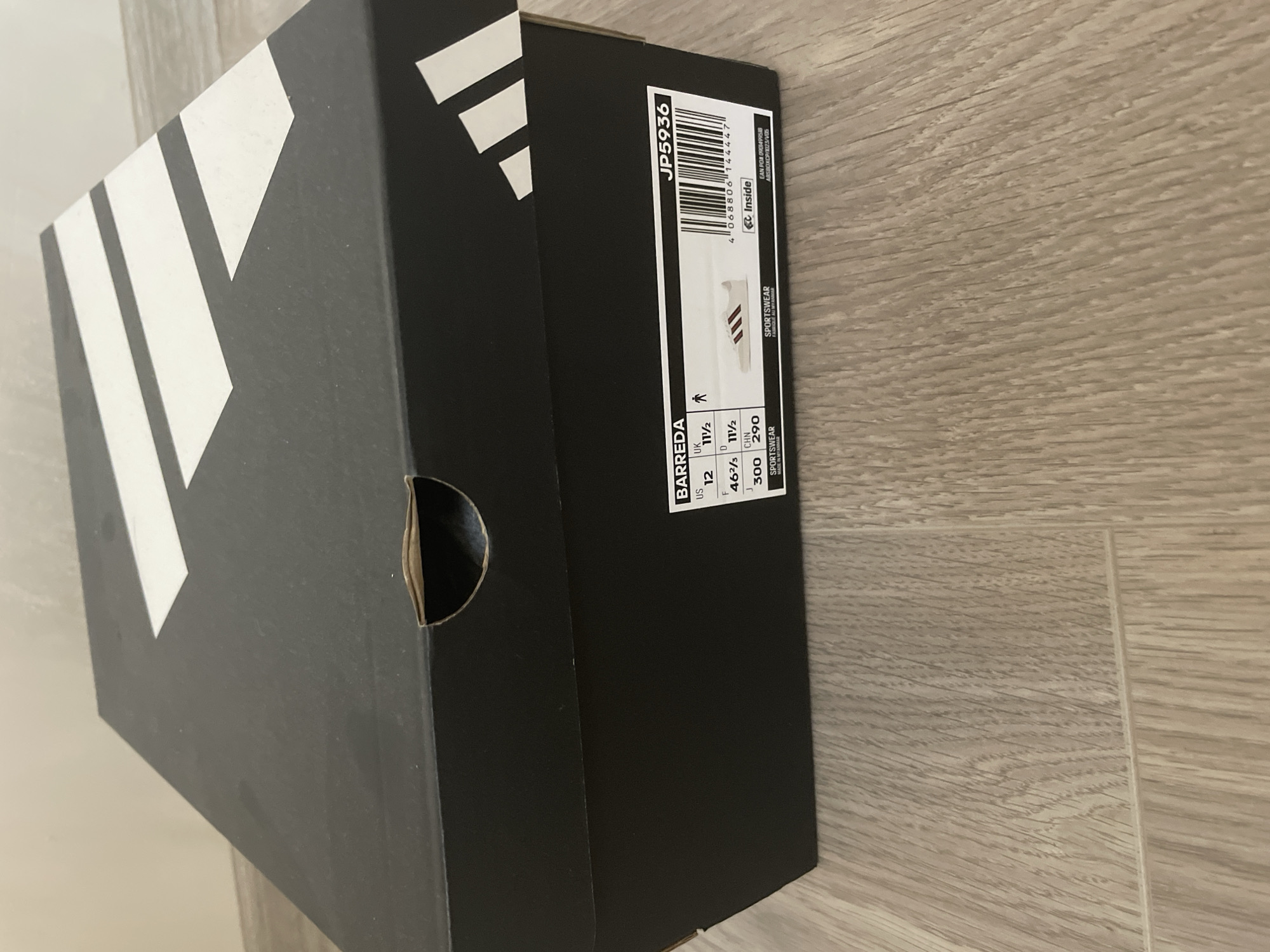 A black Adidas shoebox with white branding, featuring the iconic three stripes logo, is depicted resting on a light-colored wooden floor. The box is positioned diagonally, with its top and one side visible. A circular cut-out, likely for opening or ventilation, is present on the side of the box, showing minor wear around its edges.
A white product label is affixed to the side of the shoebox. Prominent text on the label includes the product code "JP5936" and a barcode with the number "4068806144447". The word "Inside" is visible next to a small Adidas logo. The shoe model is identified as "BARREDA". Various size measurements are listed: "US 12", "UK 11 1/2", "F 46 2/3", "D 11 1/2", "J 300", and "CHN 290". The product category is "SPORTSWEAR", and "MADE IN..." is partially visible, indicating its origin. Other alphanumeric codes include "EAN POM 0901499518" and "ABSBOXCP/023/V05". A small graphic on the label illustrates a white low-top sneaker with three dark stripes on its side.
The setting is an indoor space, possibly a home or shop in Ihtiman, Bulgaria. The floor is made of wood or laminate, characterized by light brown tones and distinct linear grain patterns. There are no people present, and no particular activity or event is occurring. The lighting is diffused, providing no specific indicators of the time of day.