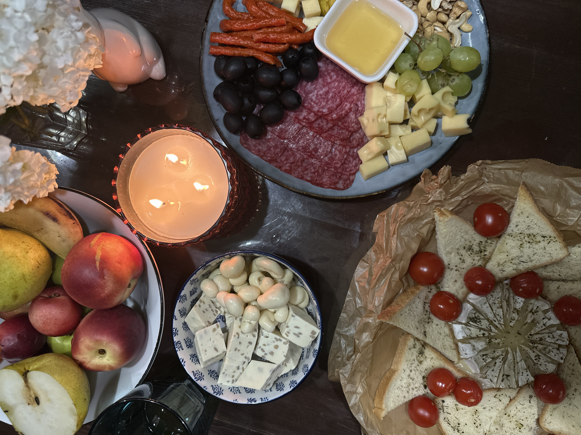 A top-down view captures a dimly lit, indoor table setting, likely an evening gathering. The scene is dominated by a collection of food items arranged on a dark wooden surface.
In the upper right quadrant, a round, blue-gray platter holds a charcuterie and cheese assortment. It features sliced salami, small red sausages, black olives, cubes of light yellow cheese, green grapes, and a small white dish filled with honey. Scattered amongst these are cashew nuts. To the left of the platter, a lit white candle in a patterned glass holder casts a warm glow. Behind the candle and to the left, a white ceramic ornament, possibly a swan or bird, sits next to a cluster of white hydrangeas.
Below the platter and candle, a white bowl contains cubes of white cheese with visible inclusions and a pile of white, bean-shaped candies. To the left of this bowl, a white dish overflows with various fruits, including a sliced pear, several red peaches or nectarines, and a few smaller red apples.
In the lower right quadrant, toasted bread triangles, sprinkled with herbs, are arranged around a round, soft cheese with a scored top. Several bright red cherry tomatoes are scattered over the bread and cheese.
The overall impression is one of a relaxed, intimate gathering, perhaps a wine and cheese tasting or a casual dinner. The warm candlelight and the abundance of food suggest a comfortable and convivial atmosphere. There is no visible text in the image.