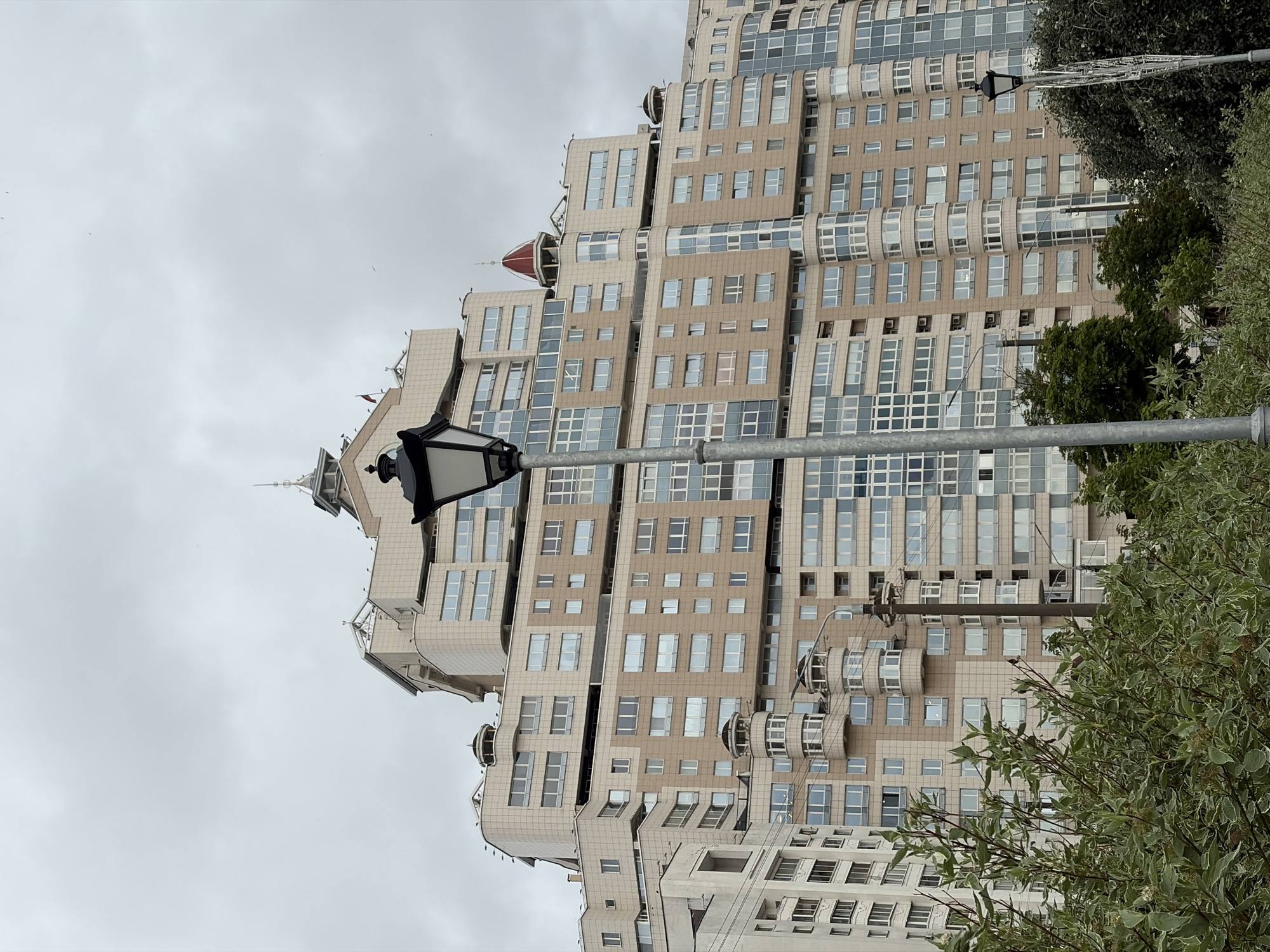 The image shows a large, multi-story residential building in Minsk, Belarus. The building is modern in design with a beige and white facade, numerous windows, and balconies. A lamppost stands in the foreground, partially obscuring the building. Trees and foliage are visible at the bottom of the frame. The sky is overcast.