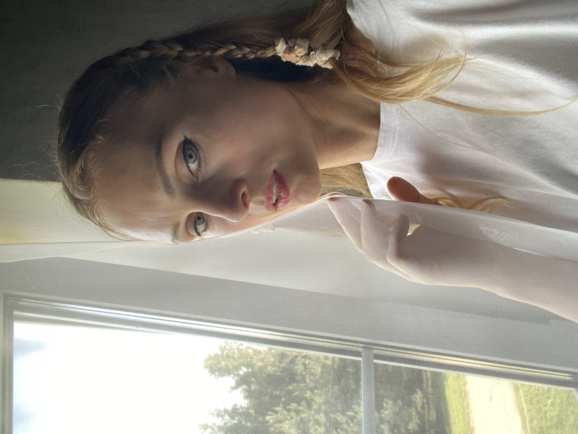A close-up, medium shot captures a woman with blonde hair braided into a single plait adorned with small, light-colored decorative elements. She is positioned in front of a window with sheer white curtains, through which trees and a green grassy area are visible. The woman is wearing a white t-shirt and her gaze is directed towards the viewer with a slight tilt of her head. Her makeup includes eyeliner and lipstick. The image appears to be taken indoors, likely in a residential setting in the area of Міханавіцкі сельскі Савет, Belarus.