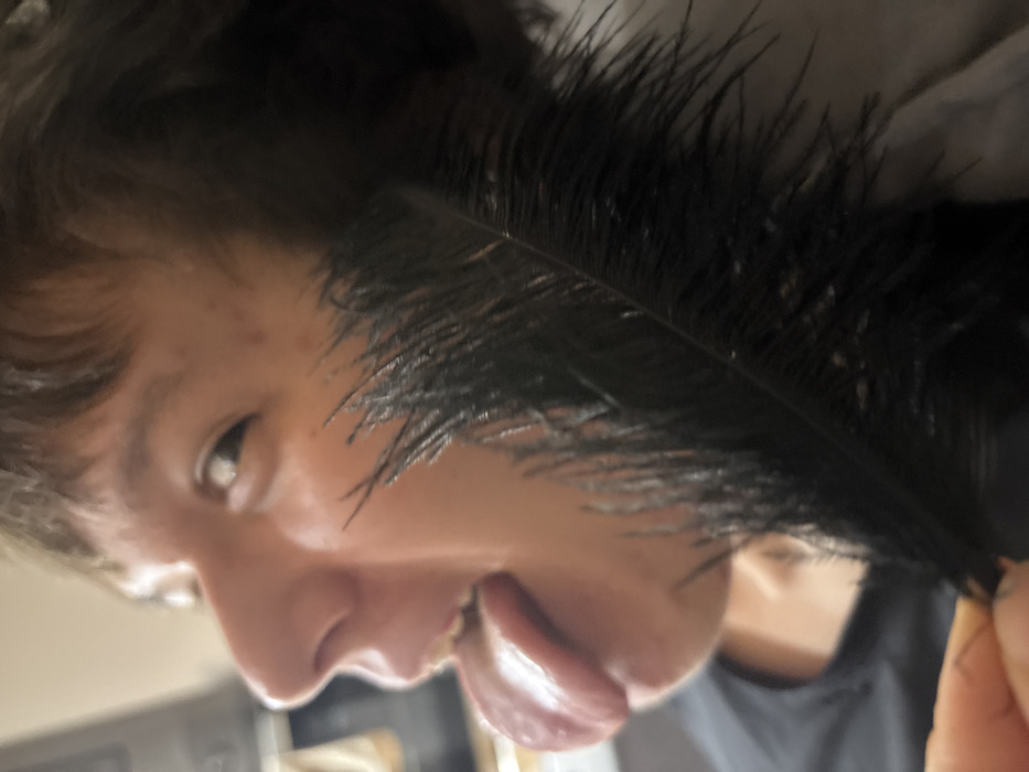 A person's face is close to the camera at an odd angle. Their tongue is sticking out and toward the camera. The person appears to be holding a large black feather close to their face. The background is blurry but appears to be an indoor kitchen setting. The image has a casual, informal feel.
