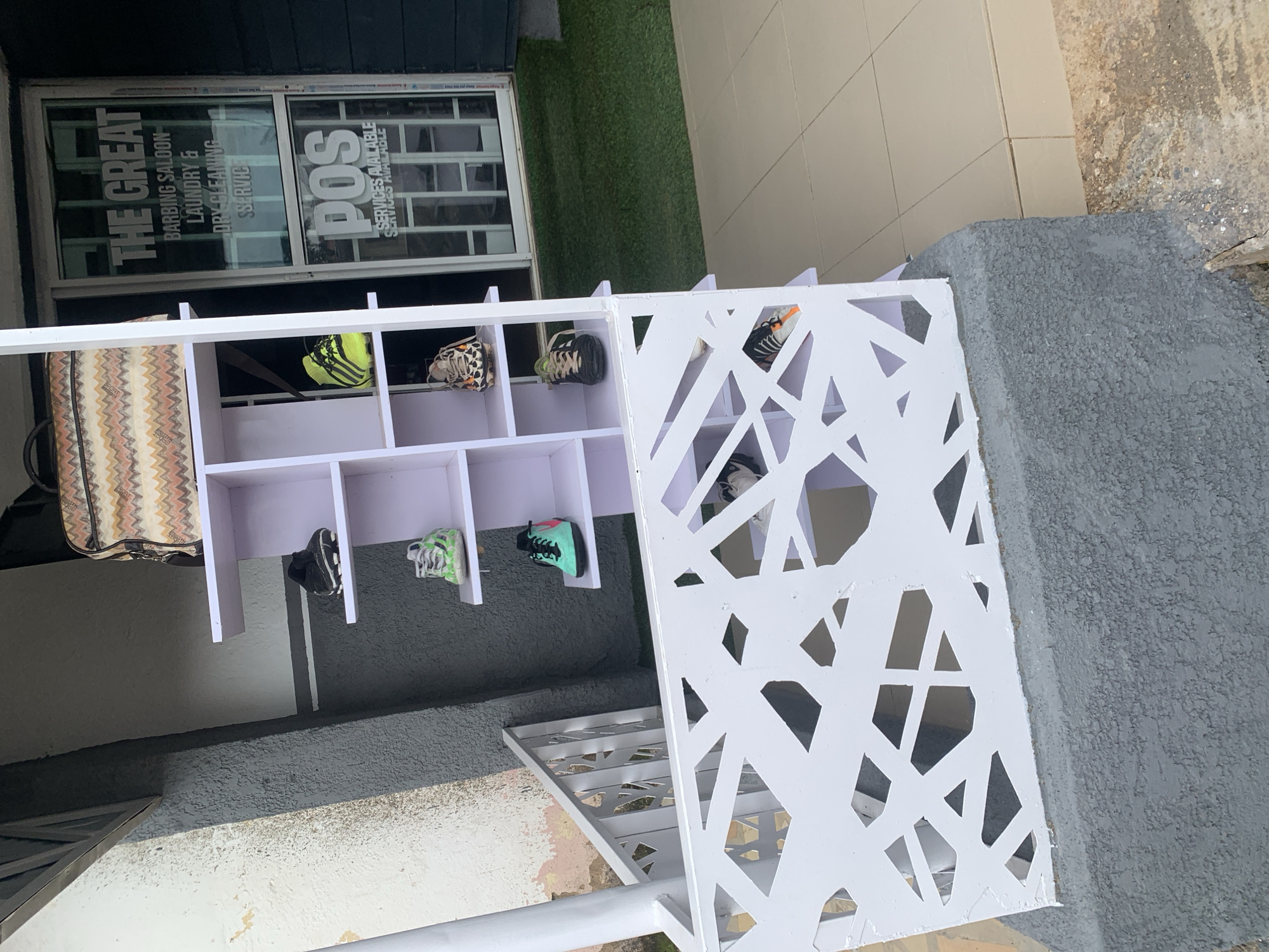 This image depicts an outdoor display of shoes outside a business in Jos, Nigeria. The shoes are arranged on a white, multi-tiered shelving unit. Some of the shoes appear to be sneakers, with various colors and designs, including a pair of bright yellow and black soccer cleats. In the background, a shop window displays text that reads "THE GREAT BARBING SALOON LAUNDRY & DRY CLEANING SERVICE" and "POS SERVICES AVAILABLE." A patterned bag rests on top of the shelving unit. The area appears to be paved with tiles and there is a patch of artificial grass to the right of the display. The setting suggests a commercial street or area within Jos.