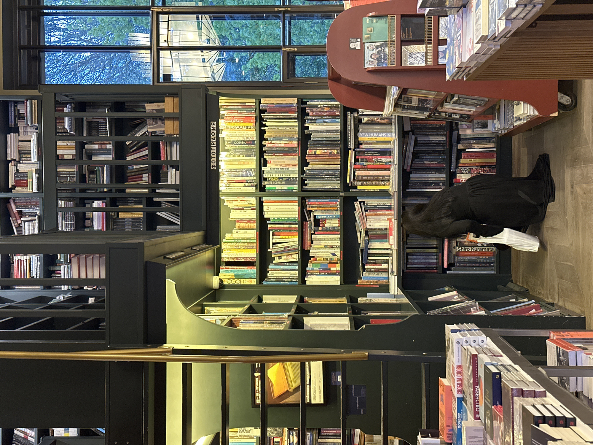 The interior of a bookstore features tall, dark green bookshelves filled with books. A person in black clothing is kneeling, presumably looking at books on a lower shelf. A rolling display table with books is visible to the right of the person. A window provides natural light above the shelves. The word "ФОТОГРАФИЯ" (Photography) is seen on the side of one shelf, and multiple books display Cyrillic text on their spines and covers.