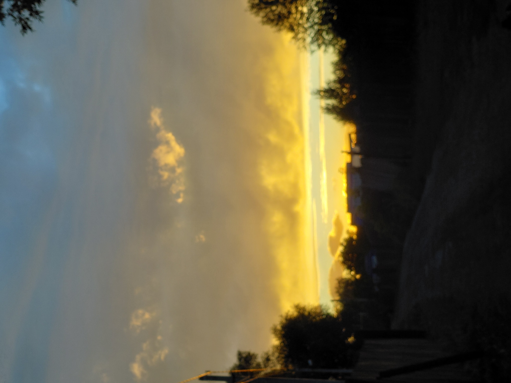 The image depicts a rural or suburban street scene at dusk, with a focus on the sky. The sky is filled with dramatic clouds, colored in hues of deep blue, gray, and vibrant yellow-orange near the horizon, indicating a sunset. There are some silhouetted trees and structures visible along the sides of a dark, unpaved road or path. A wooden fence is partially visible on the left. The overall impression is that of a quiet, dusky evening in a non-urban setting. The image's origin from Novosibirsk, Russia, suggests a likely location in one of the city's more suburban or rural districts, possibly an dacha settlement or a village on the outskirts.
