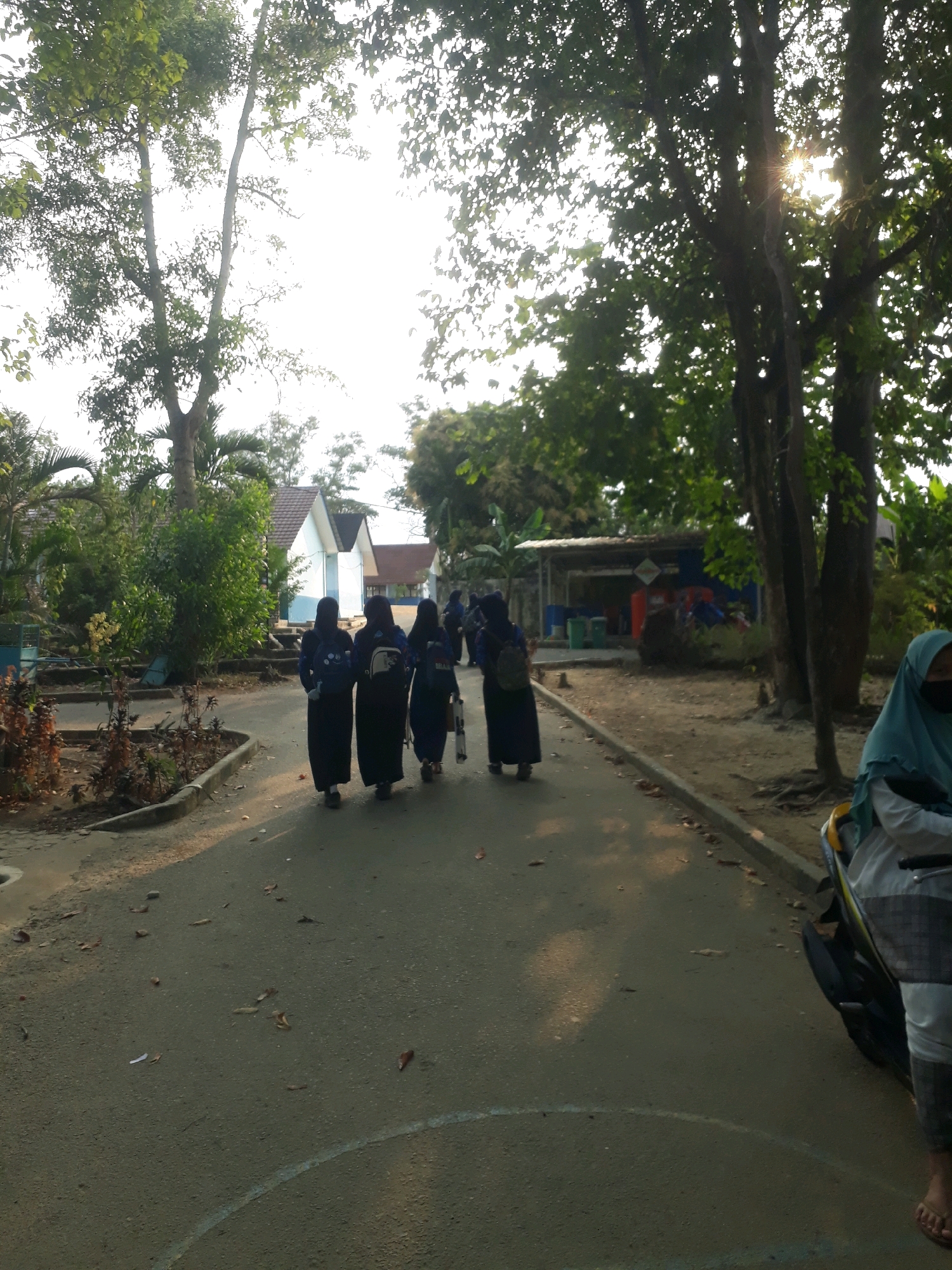 The image depicts a group of four young women, dressed in dark-colored Islamic school uniforms, walking away from the camera down a paved pathway in what appears to be a school or institutional setting. The women are wearing headscarves and long dresses, and at least two of them have backpacks. The pathway is lined with concrete curbs and some vegetation. In the background, there are several low-rise buildings with white walls and red-tiled roofs, and mature trees with dense foliage, some of which are obscuring the sky where the sun is shining through, creating a lens flare effect. The overall lighting suggests it is late afternoon. A motorcycle is partially visible on the right side of the image, with a person wearing a light blue headscarf seated on it. This scene is consistent with a school environment in Pangkalpinang, Indonesia, given the architectural style of the buildings, the vegetation, and the attire of the individuals.