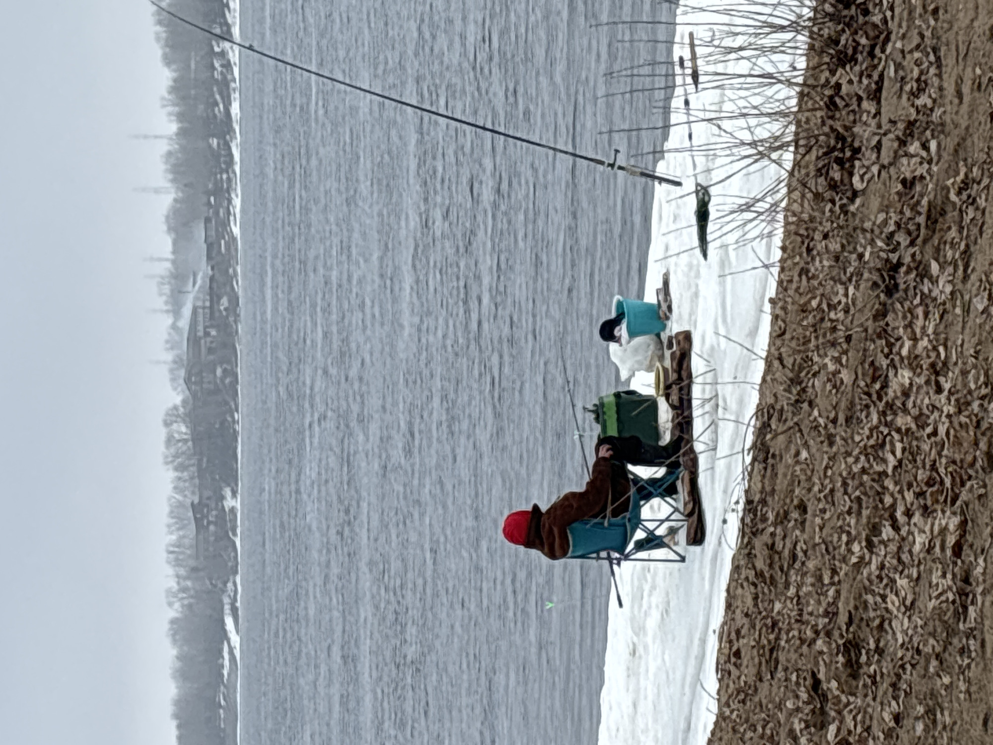 A medium shot depicts a person ice fishing on the bank of the Kama River in the city of Chaikovsky, Perm Krai, Russia. The individual, wearing a red hat and brown jacket, is seated in a blue folding chair near the edge of the riverbank. A fishing rod is set up with the line extending into the water. A blue bucket and other fishing equipment are positioned nearby on the snowy ground. The river is gray and textured, suggesting overcast weather. The opposite bank shows a line of trees, possibly with buildings visible in the distance through the bare branches. The riverbank consists of snow and frozen leaves. The overall scene suggests a cold, winter day.