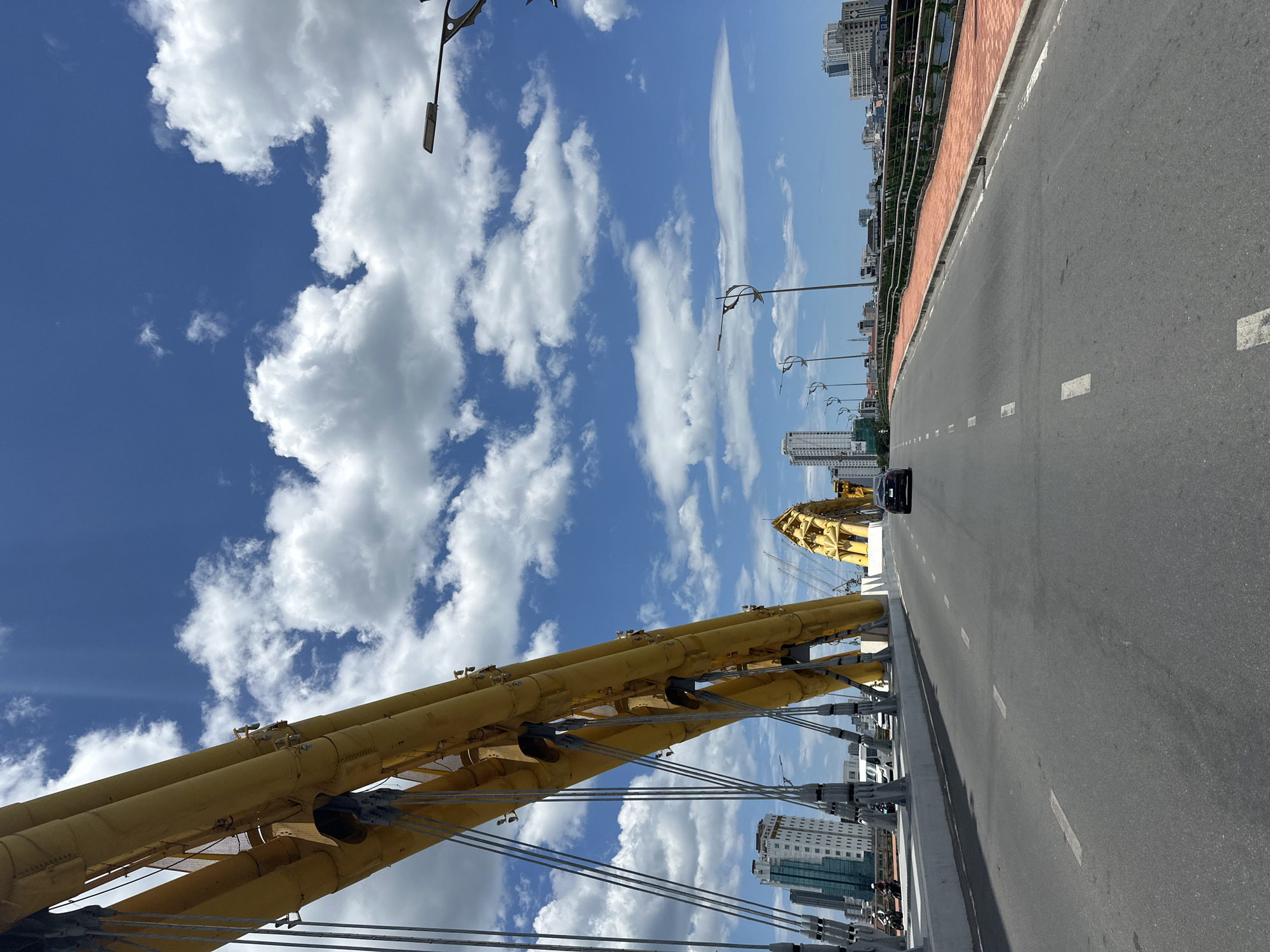 The image presents a bright, sunny daytime scene on the Dragon Bridge in Đà Nẵng, Vietnam. The perspective is taken from the bridge deck, looking along its length, emphasizing the distinctive architectural elements.
Prominently featured on the left side of the frame are large, bright yellow, tubular structural components of the bridge, resembling the body of a dragon, connected by supporting cables. To their right, a paved road stretches into the distance, marked with clear white dashed lines indicating lanes. A black car is visible driving away from the viewer on this road, suggesting moderate traffic. On the far right, a pedestrian walkway runs parallel to the road, separated by a low, orange-brown barrier.
Above the bridge, the sky is a vivid blue, filled with numerous fluffy white cumulus clouds, indicative of clear weather. In the background, on both sides of the bridge, a cityscape with various multi-story buildings and skyscrapers can be seen, hinting at an urban environment. The time of day appears to be mid-day due to the bright, direct sunlight. There is no discernible text visible in the image.