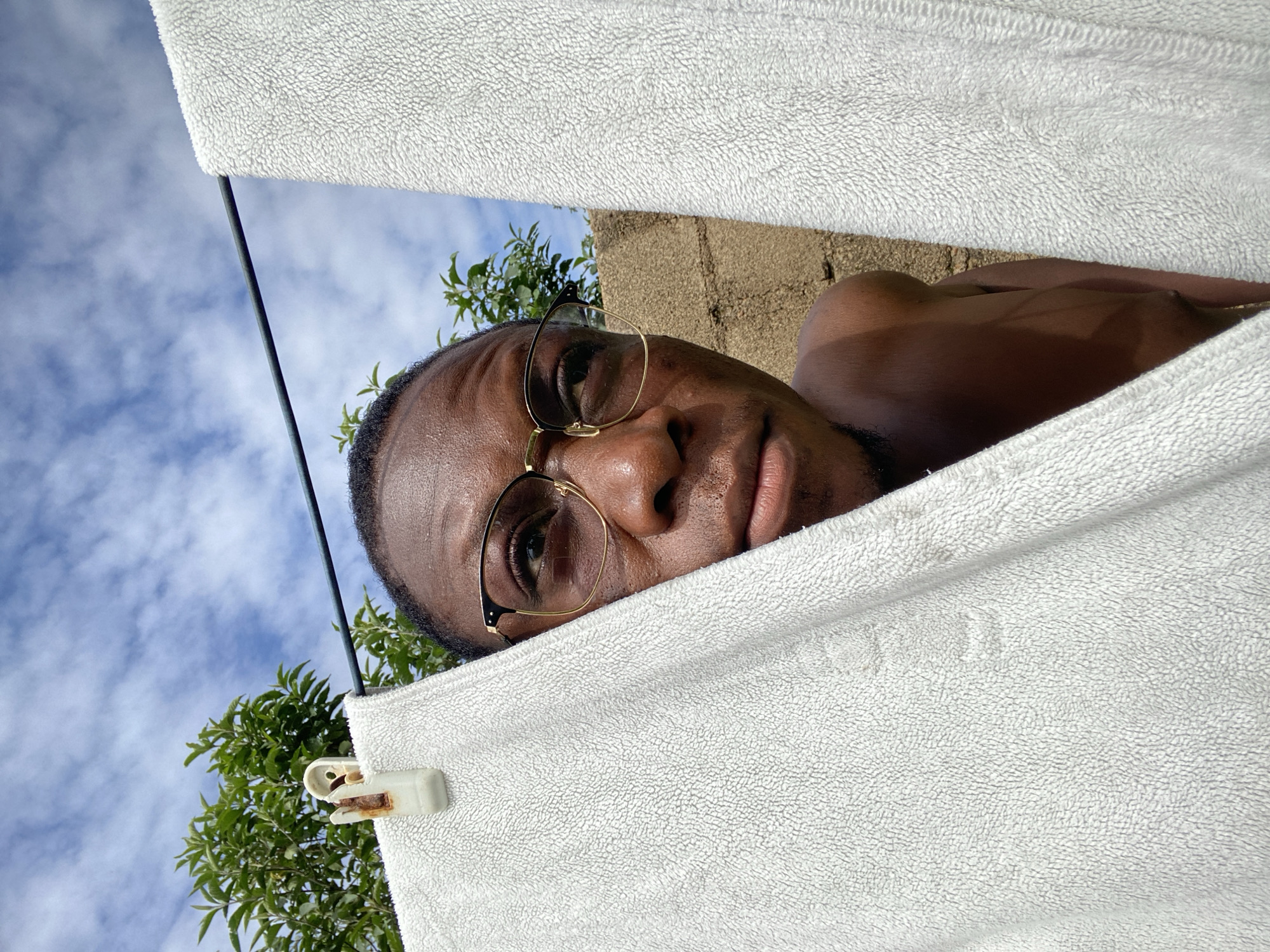 A medium shot captures a Black male wearing gold-rimmed glasses, looking upwards and to the right. He is partially obscured by two white towels hanging on a clothesline. The towel on the left is in the foreground, close to the camera, and has a white clothespin attached near the top left. The towel on the right is further back. Behind the towels, a brick wall is visible, along with green foliage and a blue sky with scattered white clouds. The image appears to have been taken outdoors in a residential area of Bukuru, Nigeria, likely on a sunny day.