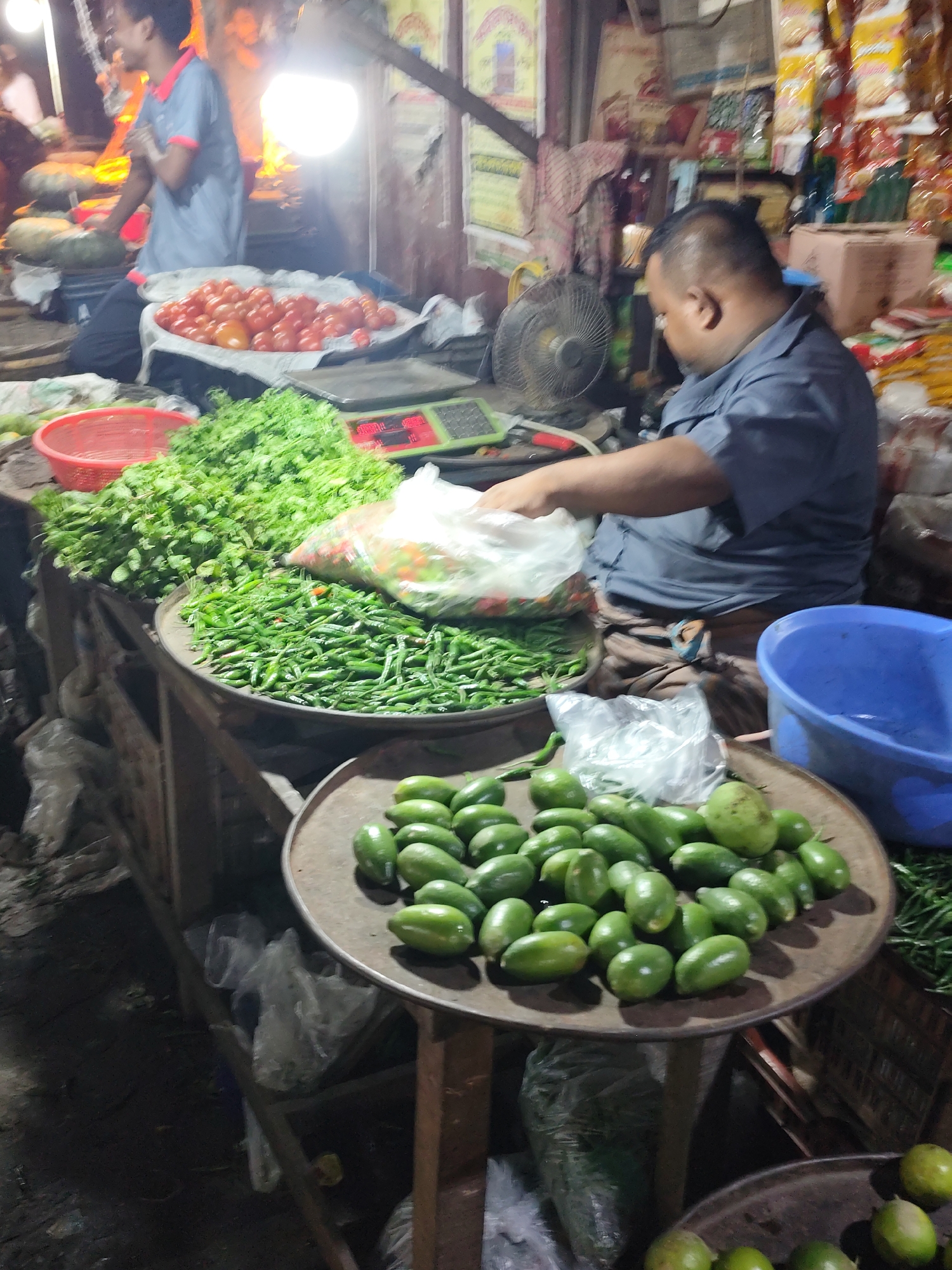 The image depicts a vibrant scene at a market in Dhaka, Bangladesh. A vendor in a blue shirt sits behind a stall, weighing vegetables. The stall displays an array of fresh produce, including tomatoes, cilantro, green chilies, and a pile of small, green vegetables that resemble baby eggplants. The area is dimly lit, but a bright light illuminates the upper section of the stall. A young man in a blue shirt is visible in the background near the tomatoes, interacting with someone. The market appears bustling, with various unidentified objects and packages stacked around the vendor and stall. A small electric fan is positioned to cool the vendor.
