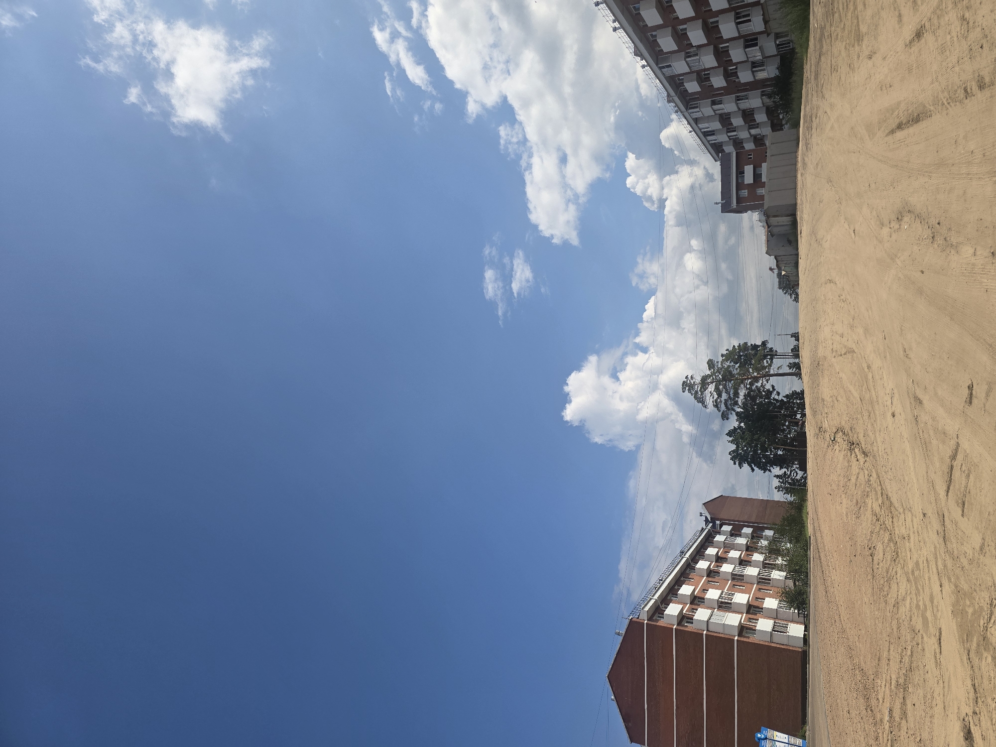 The image depicts a rural or semi-urban landscape under a bright blue sky with scattered white clouds. In the foreground, a sandy, unpaved road or path slopes upwards. To the left, a multi-story brick building with white-trimmed balconies is visible. Further down the road, more buildings and a cluster of pine trees are present. The right side of the image shows another similar multi-story building. The scene appears to be a construction site or an underdeveloped area. The location is specified as the city of Ulan-Ude, Russia. There are no people or animals in the image.