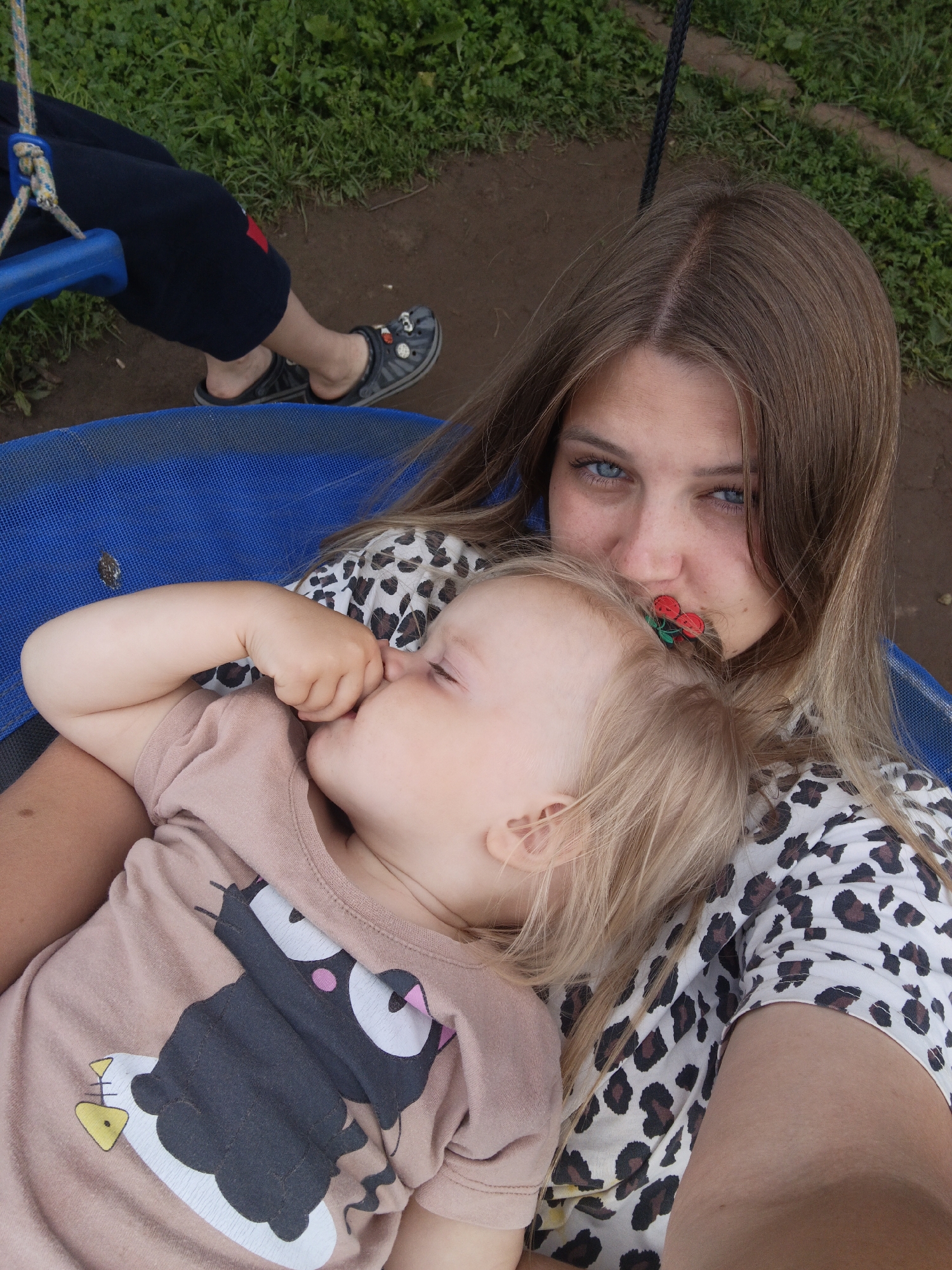 A selfie is taken by a young woman with long blonde hair and blue eyes. She is holding a sleeping toddler, who is resting on her chest. The toddler has blonde hair and is wearing a light brown t-shirt with a cartoon cat design. The woman is wearing a white shirt with a leopard print. A red and green hair clip is visible on the woman's hair. They are both sitting on a blue swing. In the background, there is green grass and brown dirt. The image appears to be taken outdoors in a rural setting, possibly in Medvedyovka, Russia.