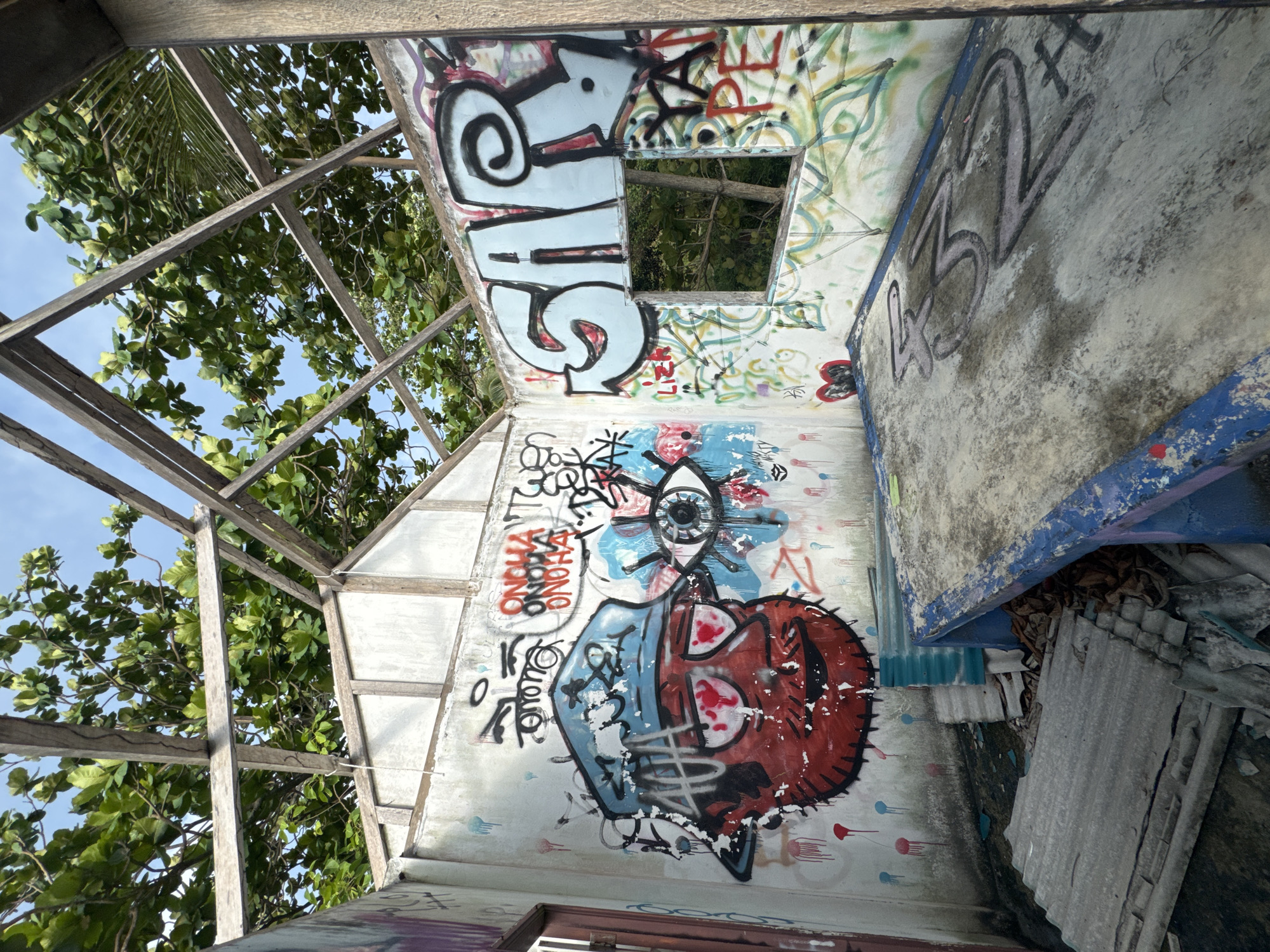 The image depicts the interior of an abandoned structure in Baan Thong Nang, Thailand, possibly a derelict building or shelter, based on the incomplete wooden frame visible through a partially missing roof. The walls are heavily covered with graffiti, including stylized lettering, abstract designs, and figurative elements like a stylized eye and a red heart. The perspective is from within the structure, looking upwards towards the sky, which is visible through the open roof. Debris and rubble are present in the lower right corner of the image, further suggesting the building's state of disrepair. The presence of vegetation (trees) is visible through the roof and around the wooden frame, indicating the building is exposed to the natural environment.