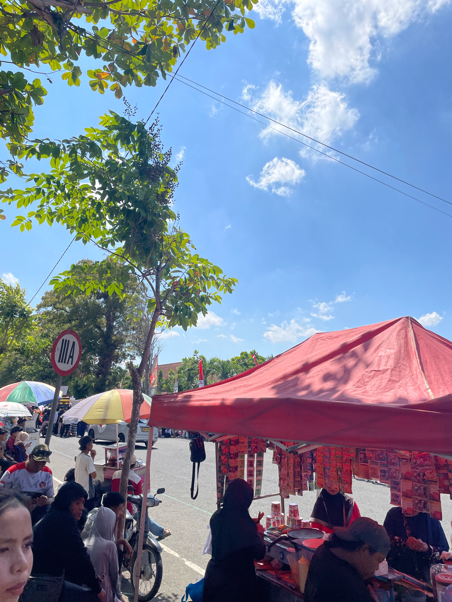 The image captures a bustling street scene in Purworejo, Indonesia, on a bright, sunny day. A red canopy tent, characteristic of roadside stalls, dominates the foreground, displaying numerous small packets hanging from strings, likely food or merchandise. Underneath the tent, several individuals are engaged in activity, possibly preparing or selling goods from a grill and counter setup. One person in a black hijab and red and black patterned clothing appears to be serving customers.
Beyond the stall, the street is lined with more vendors and shoppers. Colorful umbrellas, green, white, and yellow, shelter some of the other stalls. People are milling about, some sitting, some standing, and a few are on motorcycles. A motorcycle is parked near the red tent, with several people seated on it.
In the background, trees provide shade, and a round traffic sign with white and red markings, displaying the Roman numeral "IIIA," is visible. Power lines crisscross the bright blue sky, which is dotted with fluffy white clouds. The overall impression is one of a lively local market or street fair in Purworejo.