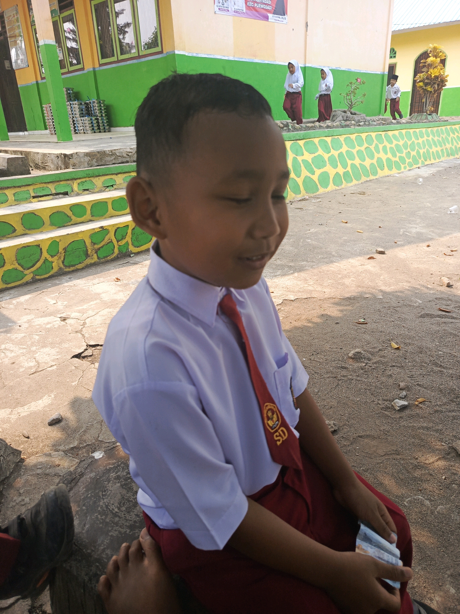 An outdoor medium shot captures a young boy wearing a white school shirt with a red tie and maroon pants, sitting on a concrete step. The boy is looking to his right with his eyes partially closed and a slight smile on his face. He is holding a stack of folded currency in his hands. In the background, a school building with yellow and green accents is visible, along with other students in similar school uniforms walking in a courtyard. The ground is paved with concrete and some gravel. The environment appears to be a school in Karya Dadi, Indonesia, suggested by signage in the background.