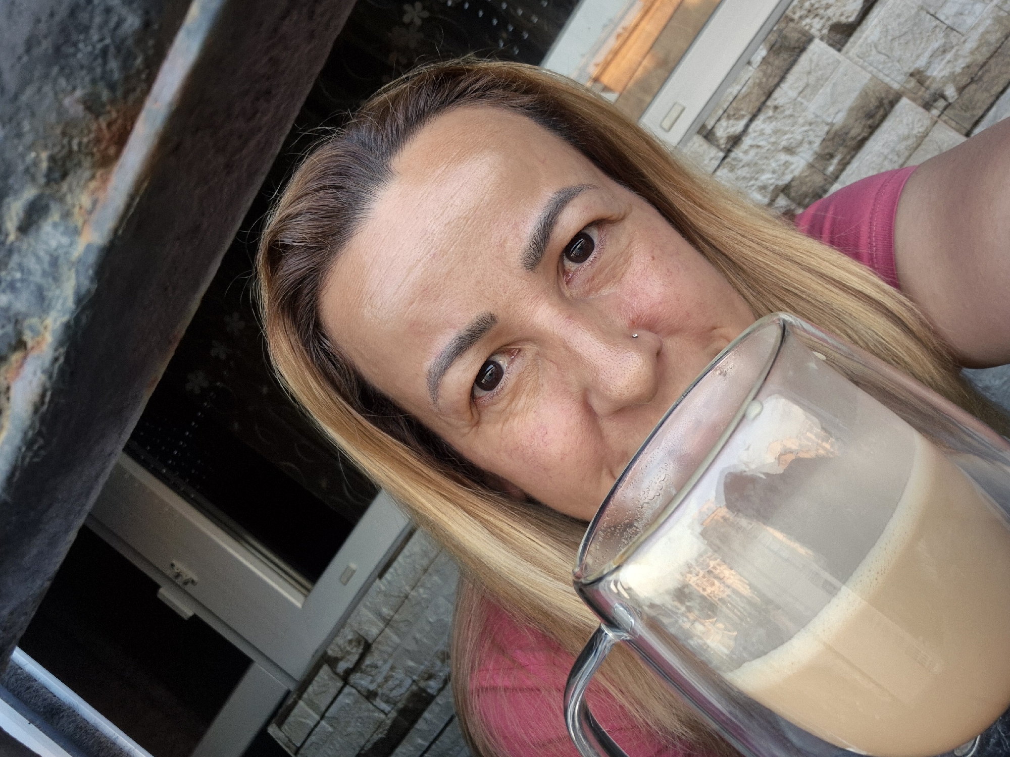 A medium shot captures a person with light brown hair and fair skin drinking from a transparent mug of coffee. The person is wearing a pink shirt and has a nose piercing. The background shows a textured wall, a window, and a dark, possibly metallic, overhead structure. The scene appears to be outdoors or near a window with natural light. The camera angle is a close-up, looking upwards towards the person and the mug.