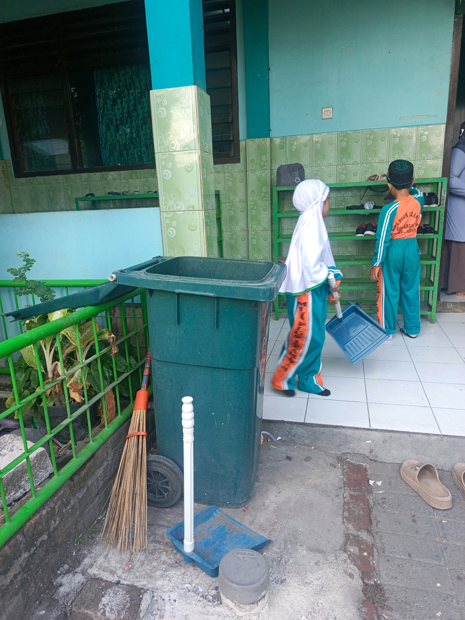 The image depicts an outdoor scene near a building in Banjarsari, Indonesia, likely a school. Two children, dressed in matching green and orange uniforms with headscarves, are the main subjects. The child on the left, facing away from the camera and wearing a white headscarf, is holding a blue dustpan. The child on the right, also facing away, is wearing a dark cap and stands beside a green shoe rack filled with footwear. In the foreground, a large green trash bin is positioned next to a broom with an orange handle. The ground is paved with light-colored tiles. A green railing and potted plants are visible on the left side of the frame. The building's exterior features teal-colored walls and green tiled lower sections.