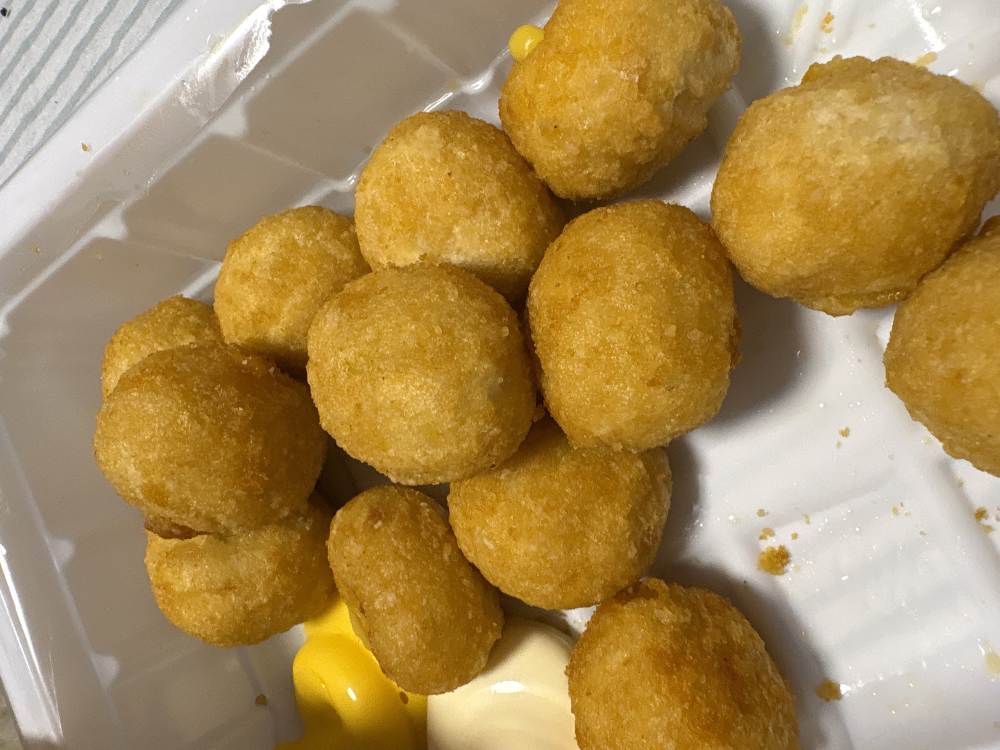 The image shows a white plastic tray filled with approximately fifteen golden-brown, spherical objects, likely fried potato or cheese balls. There are dollops of yellow and white sauce at the bottom of the tray. The lighting is bright, suggesting an indoor setting. There are no people or indications of a specific location mentioned in the image. The location of Прикамский, Russia is not related to the image.
