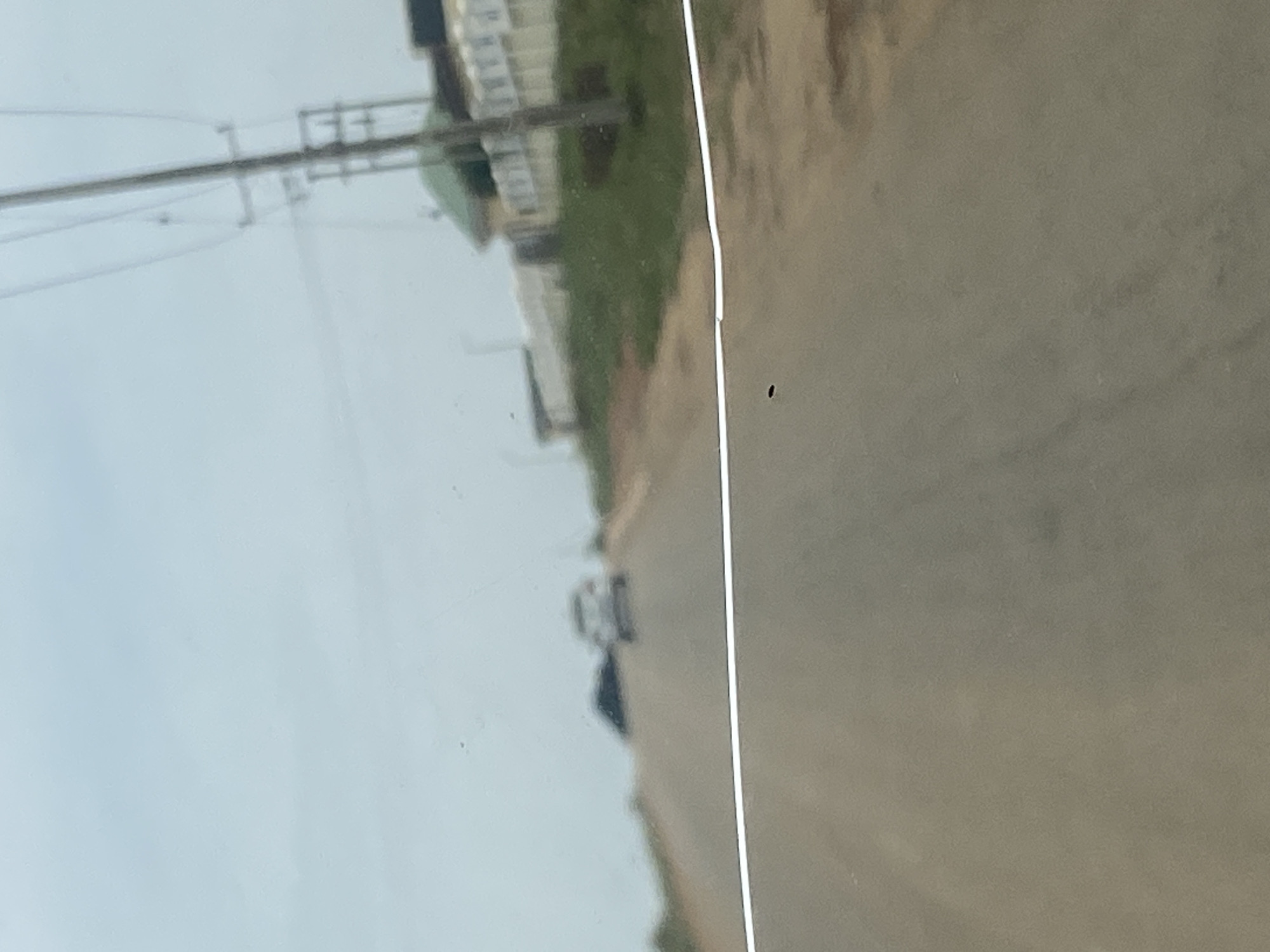 The image depicts a view from a moving vehicle on a paved road, seemingly in a semi-urban or rural area outside the city of DU, Nigeria. The road stretches uphill and curves slightly to the right. There are two cars visible on the road ahead, both slightly blurred due to motion. The car on the left is dark and appears to be a sedan, while the car on the right is lighter in color and also appears to be a sedan. The environment on the sides of the road consists of dry, brown earth with sparse green vegetation. In the background, on the right side of the road, there are buildings with white walls and green roofs, possibly residential or institutional structures, bordered by a fence. A utility pole with wires is also prominently visible on the right. The sky is overcast and a light blue-gray. A thin, white line, likely a reflection or a scratch on the vehicle's windshield, runs horizontally across the lower part of the image. A small, dark, oval-shaped object, possibly an insect, is visible on the road surface in the lower center of the image. The overall impression is a mundane journey through a developing area.