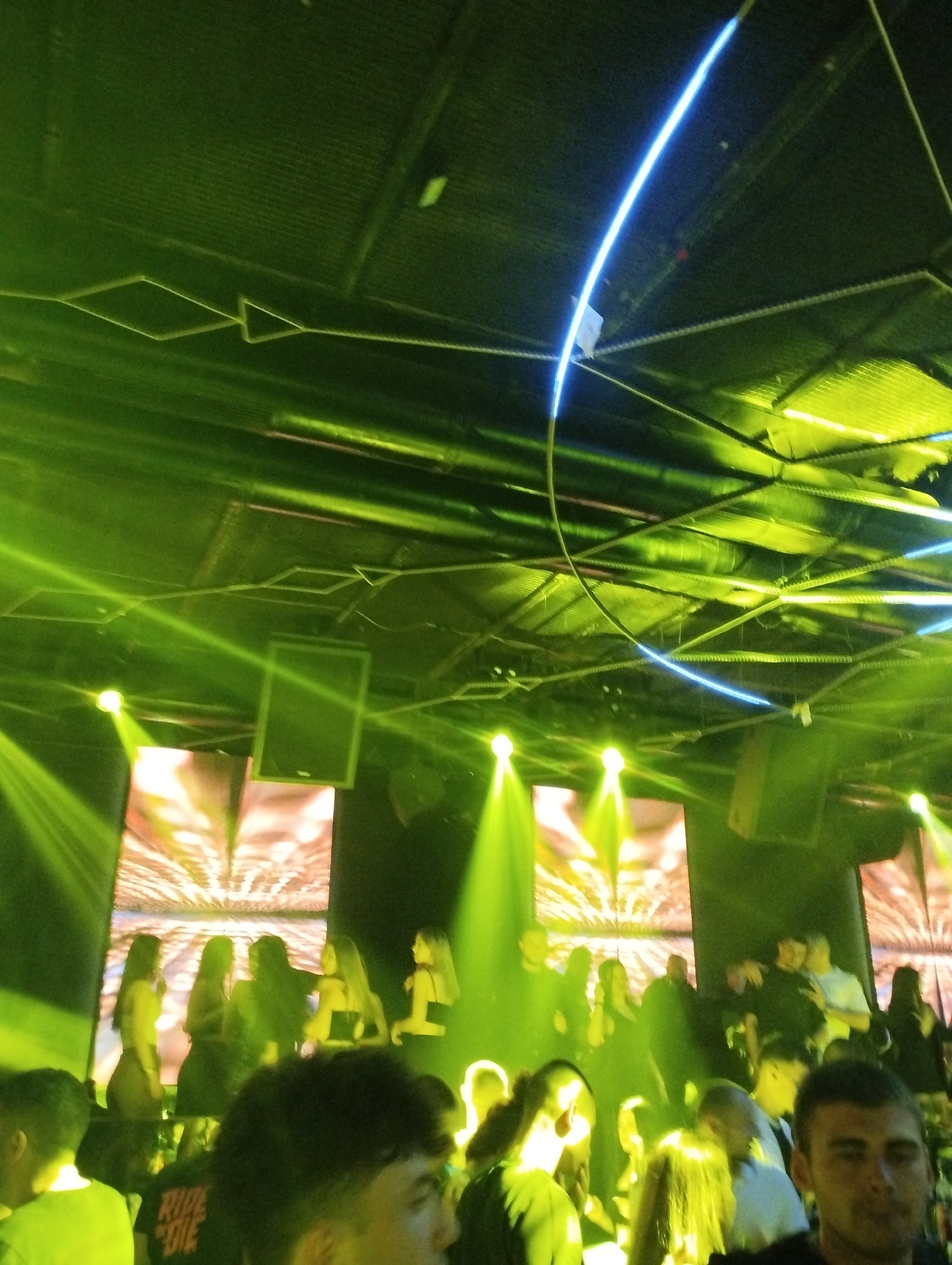 The image shows a dimly lit, crowded indoor space, likely a nightclub or bar in Unknown. The lighting is a combination of green spotlights and a blue neon tube suspended from the ceiling. There are several people in the foreground and middle ground, mostly facing away from the camera. Some appear to be standing, talking, or dancing. In the background, there are illuminated screens or panels with abstract patterns. There are also loudspeakers mounted near the ceiling.